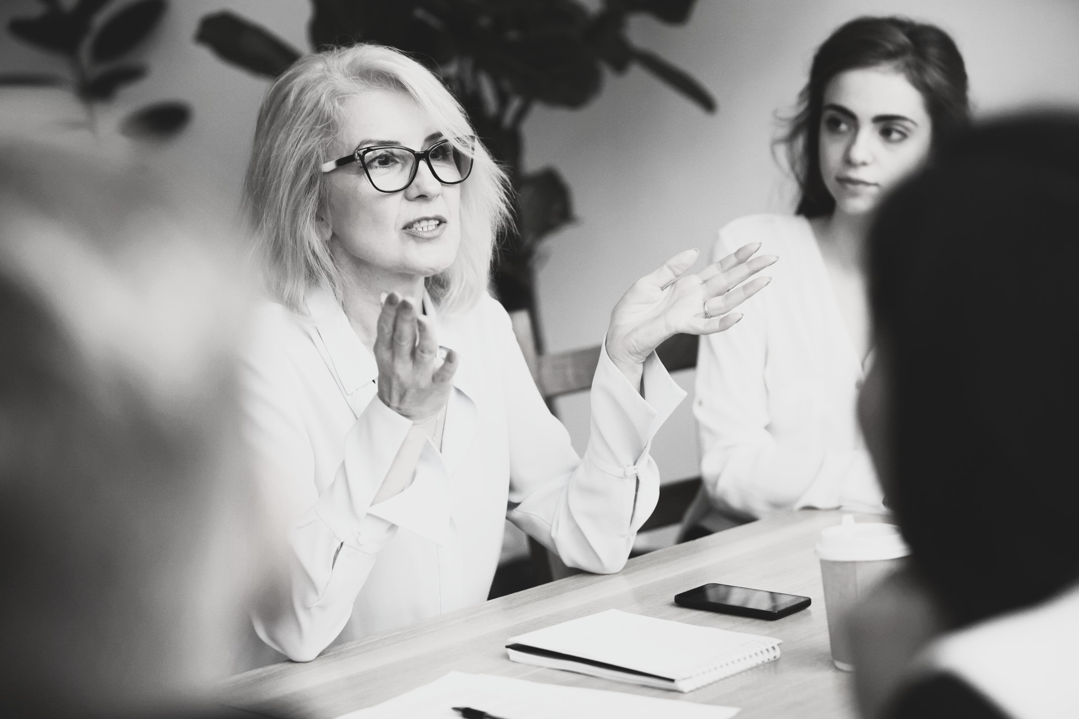a woman in a white shirt and glasses sitting at a meeting table gesturing with her hands