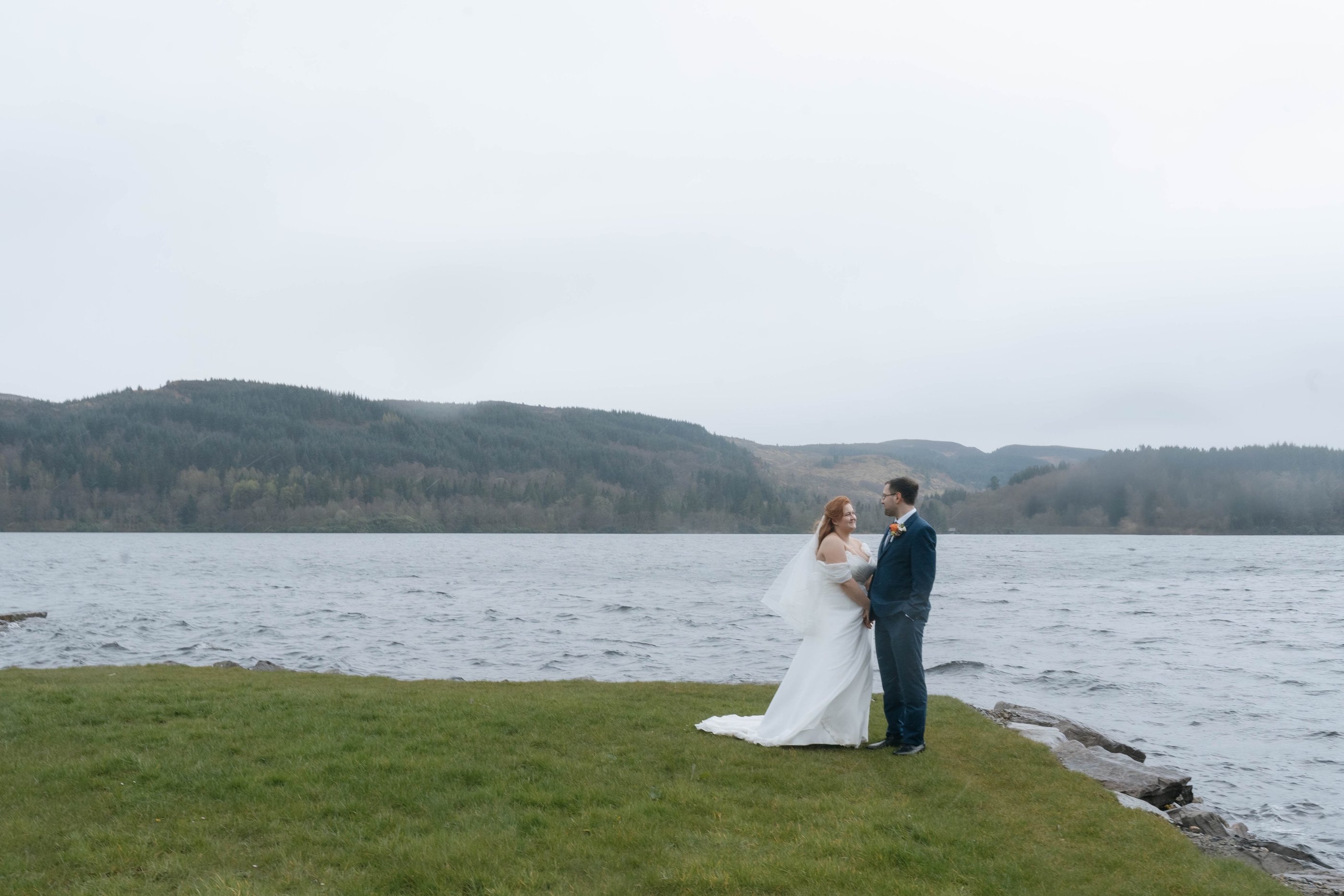 A bride and groom posing for wedding photos by a scenic lake in Scotland on a misty day.