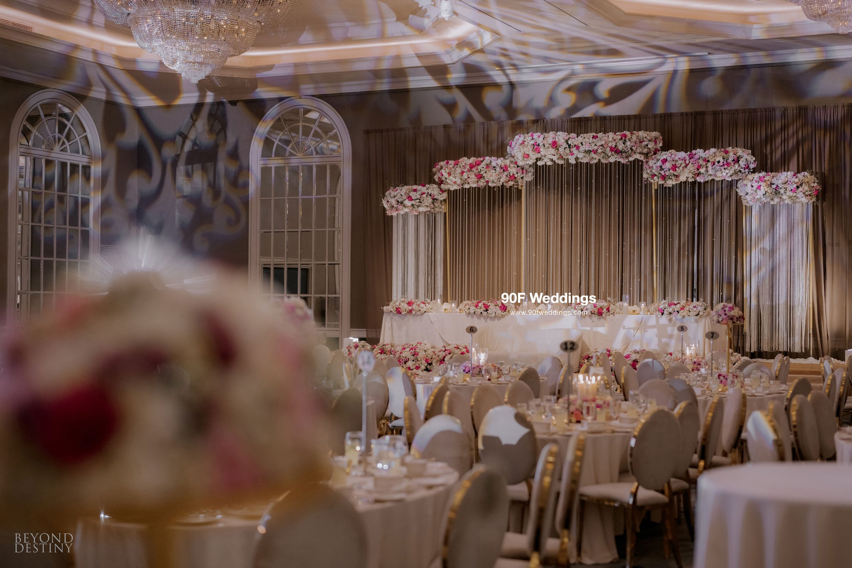 Elegant luxury wedding ballroom reception featuring a floral stage backdrop and crystal chandeliers.