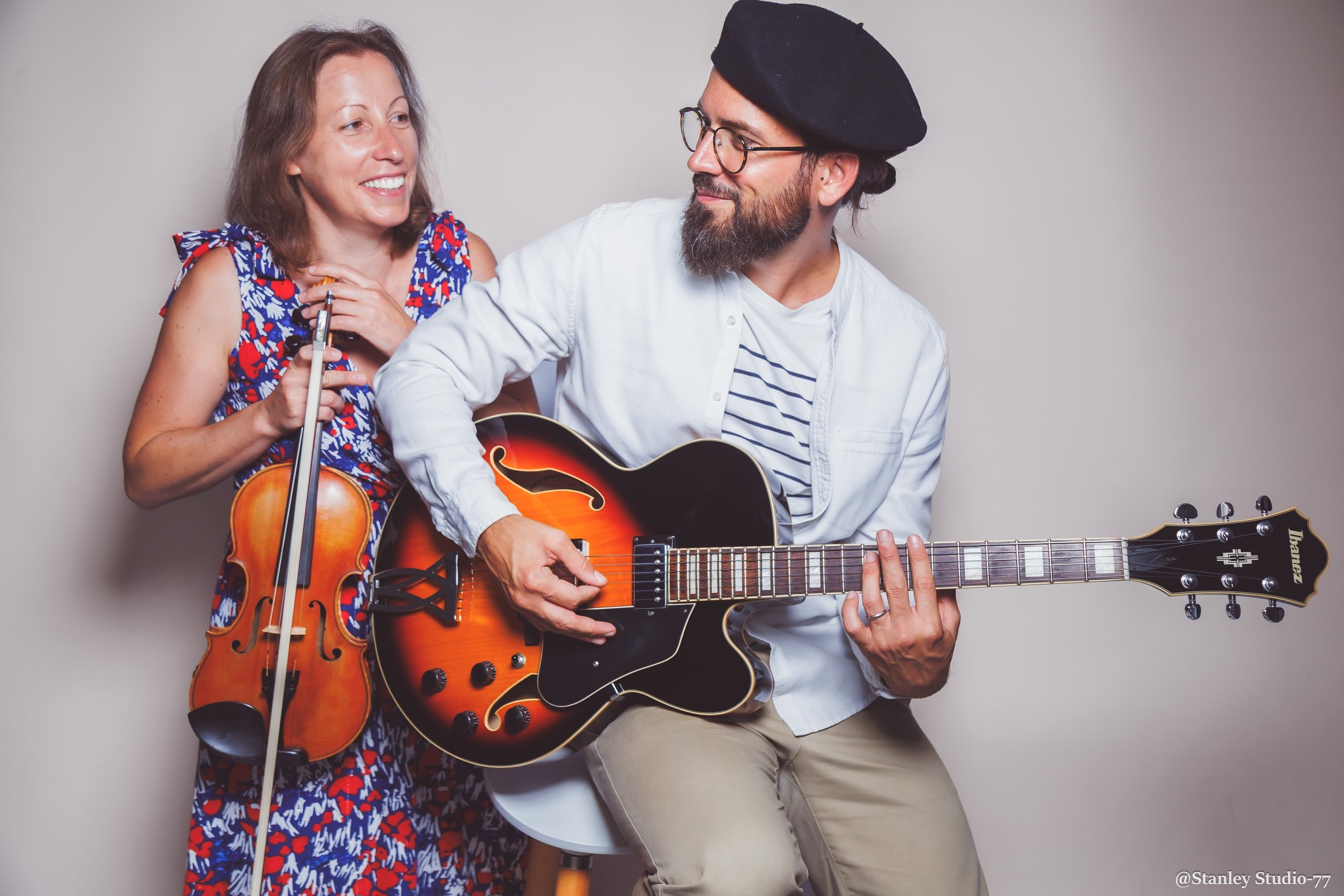A smiling man plays a sunburst hollow body electric guitar next to a female violinist.