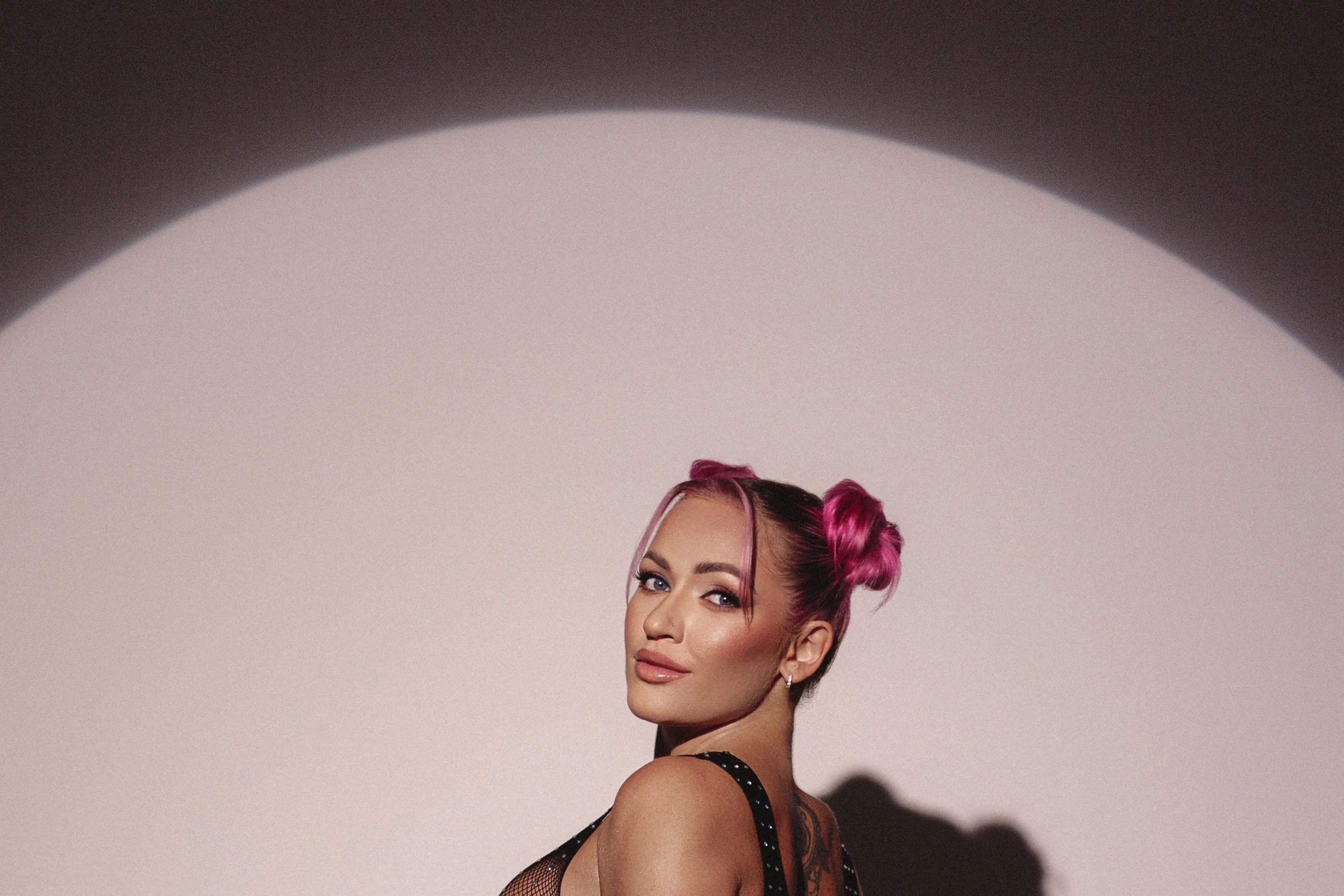 A stylish woman with pink space buns hair posing in a circular spotlight against a dark background.