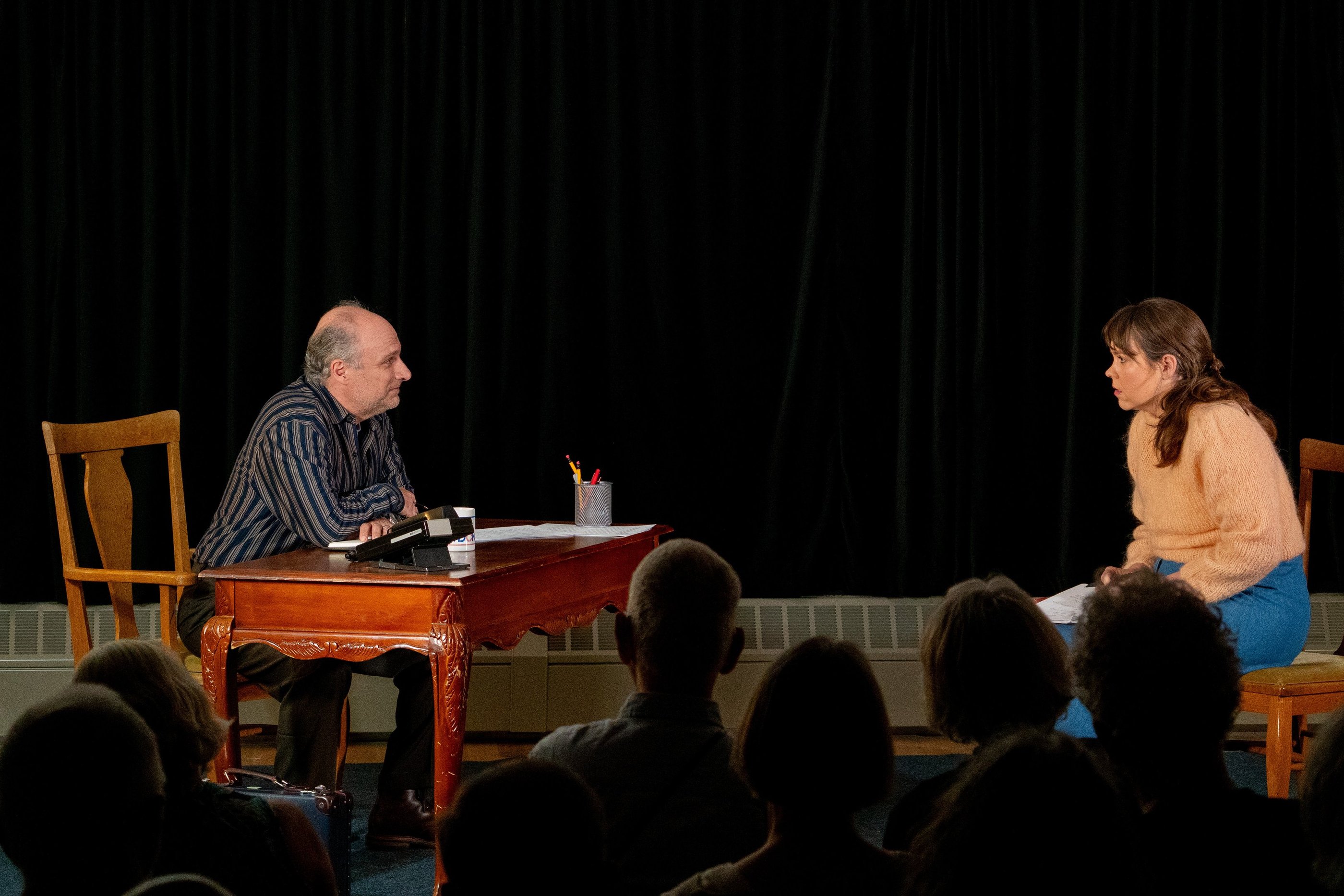 Man seated at desk across from woman seated in a chair. They are in front of an audience