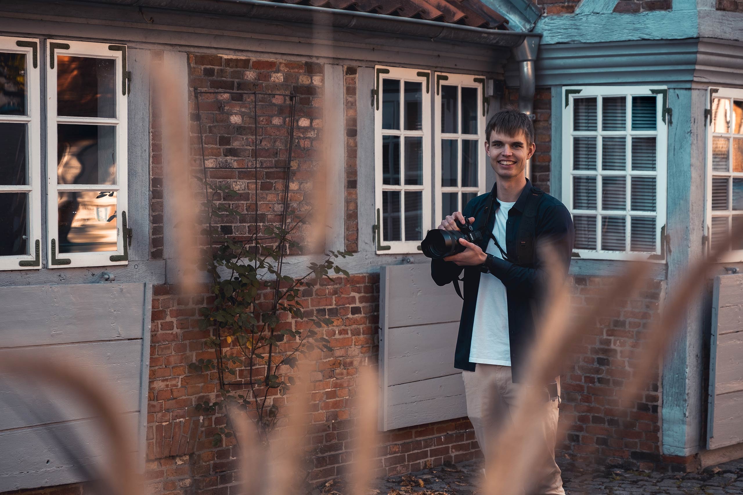 Lächelnder Fotograf Joel Pingel steht mit Kamera vor einem Fachwerkhaus. Portrait von Just Perspectives.