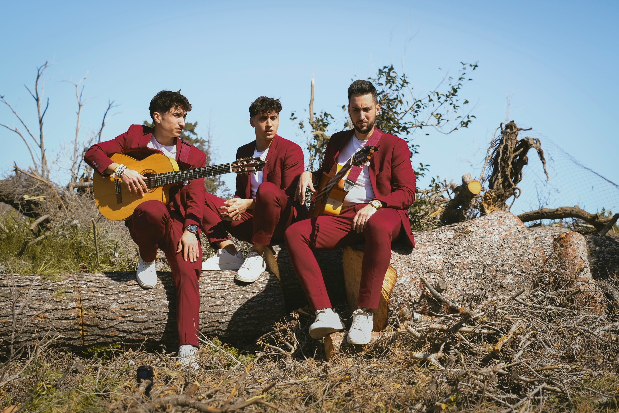 Three male musicians in burgundy suits playing acoustic guitars while sitting on a large log outdoors.