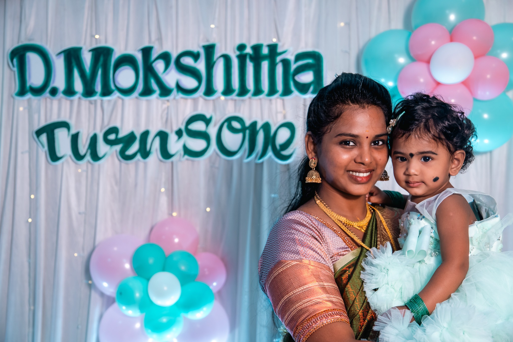 A mother holding her baby daughter at D. Mokshitha's first birthday event photographed by Memories a