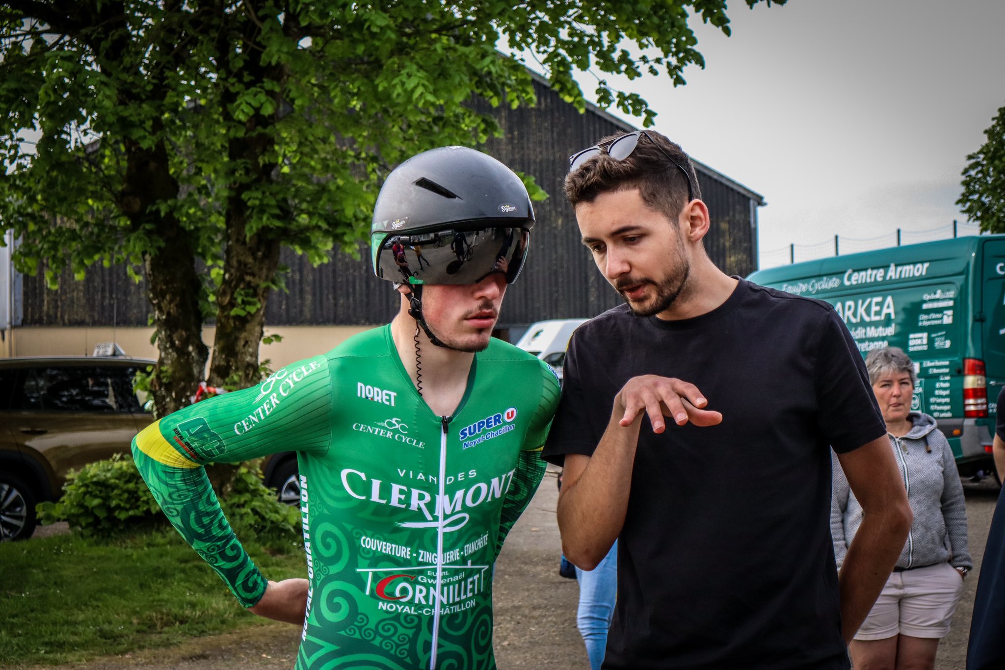 Professional cyclist in green team jersey and aero helmet discussing race strategy with his coach.