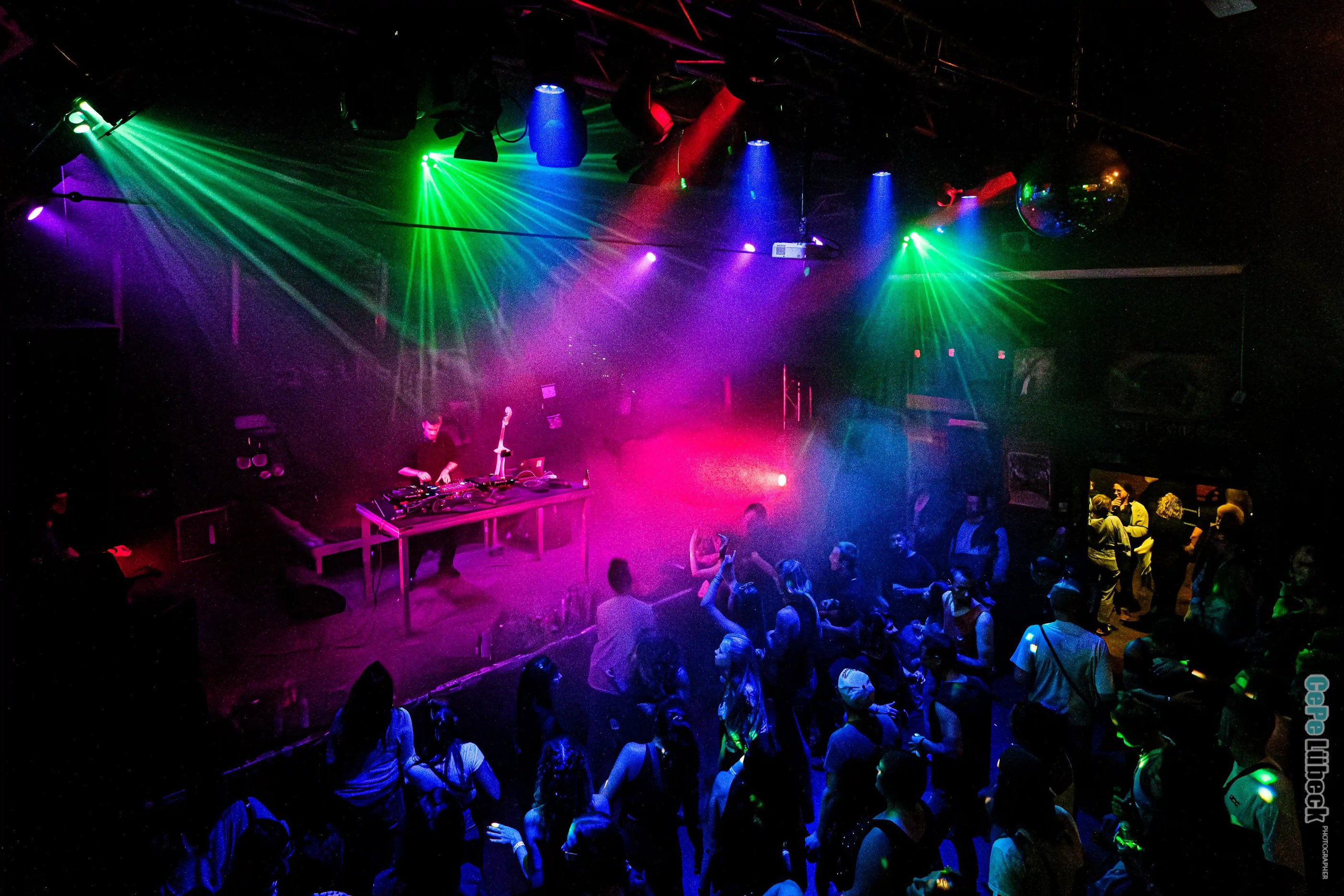 A DJ performs on a dark stage with vibrant neon laser lights and a dancing crowd at a nightclub event.