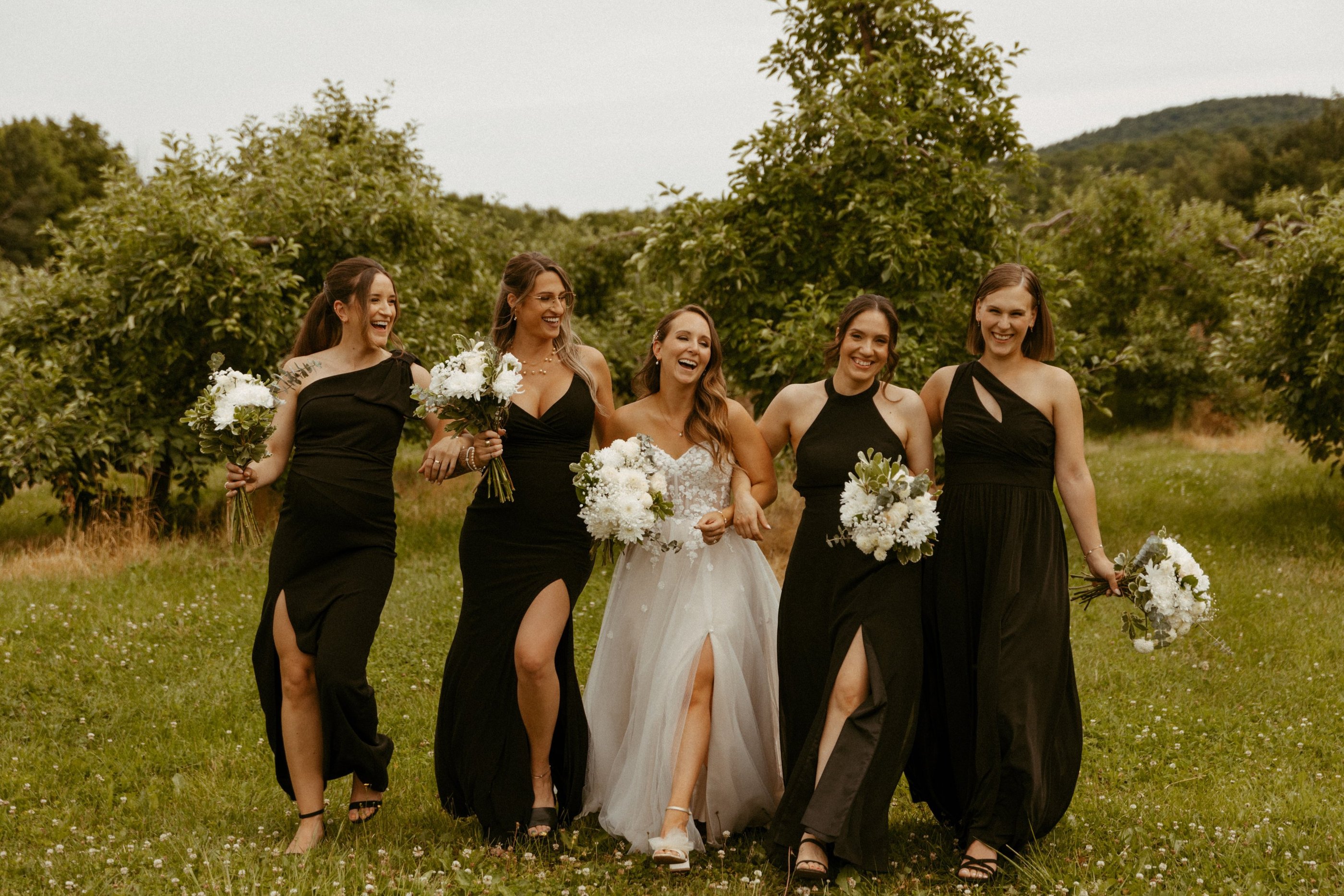 A bride in a lace gown walks and laughs with bridesmaids in black dresses at an outdoor wedding.