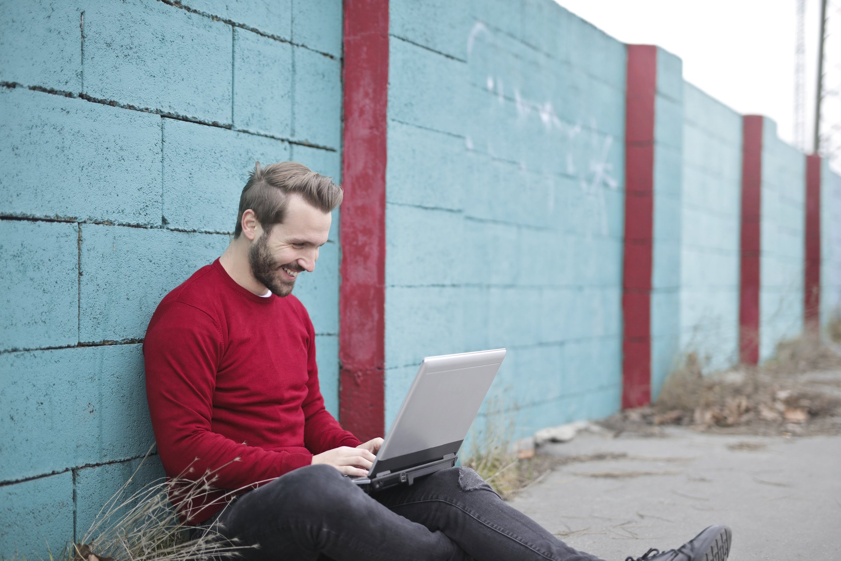 A man sits against a wall using a laptop. He is wearing a red jumper and is smiling.