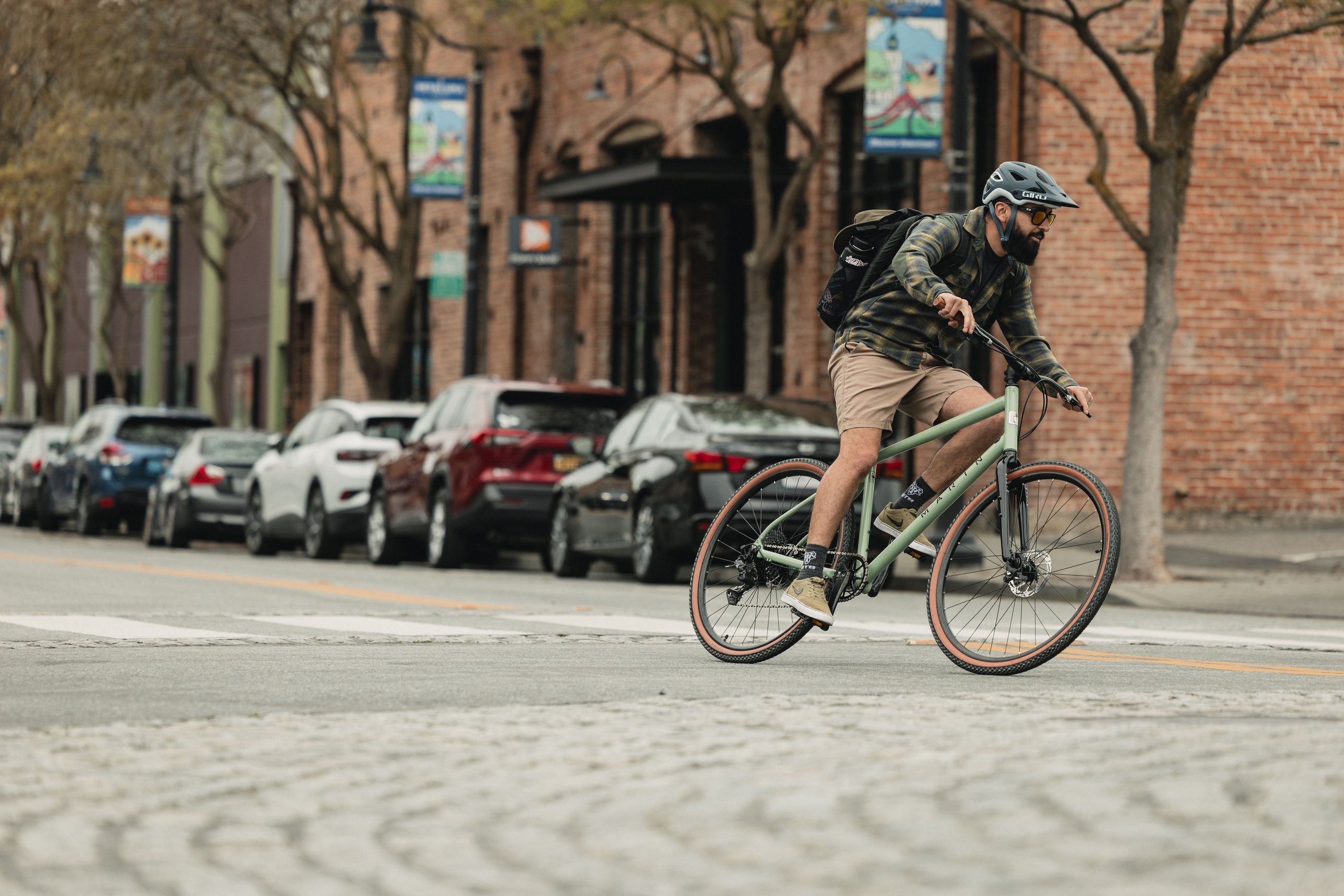 A male cyclist wearing a helmet and backpack rides a gravel bike through a city street.