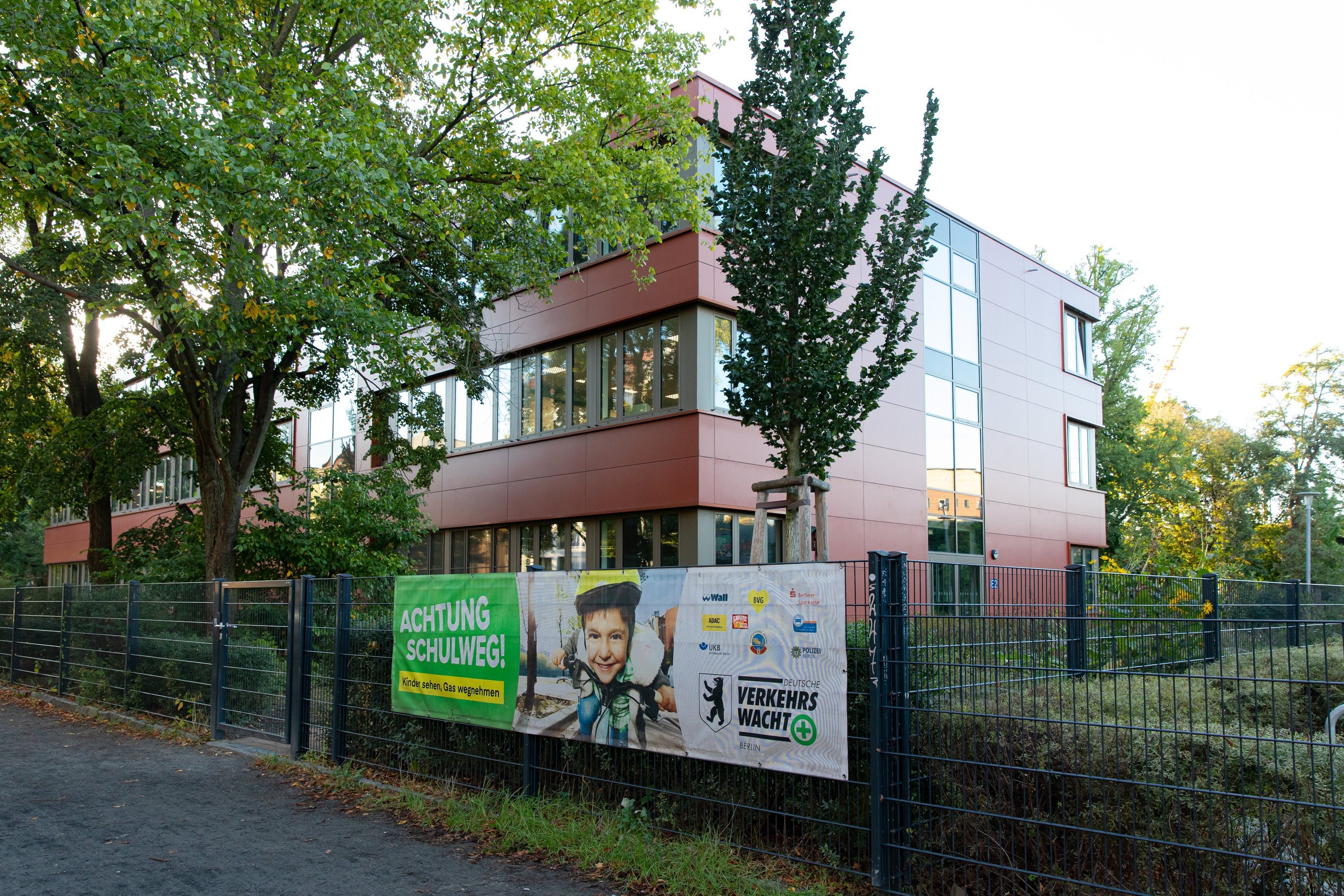 Foto von einerm roten Schulgebäude, an dem Zaun davor ist ein Plakat zur Schulwegsicherheit.
