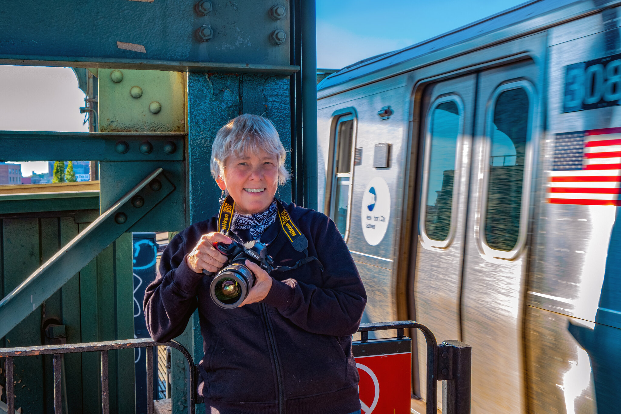 A fotógrafa Martha Cooper, segurando uma câmera e sorrindo, posa em um ambiente urbano