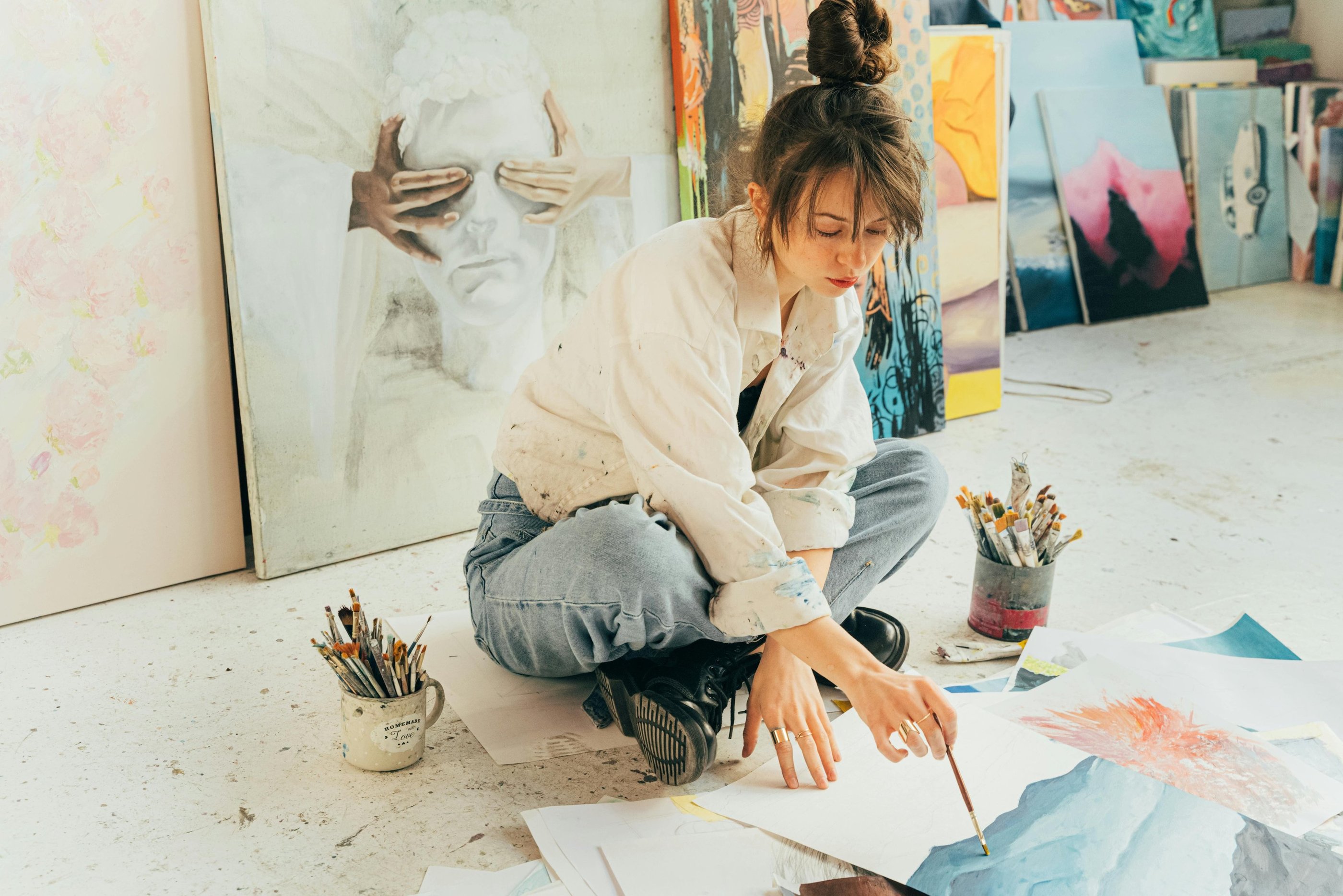 Jovem artista sentada no chão rodeada por pinturas, trabalhando em uma aquarela.