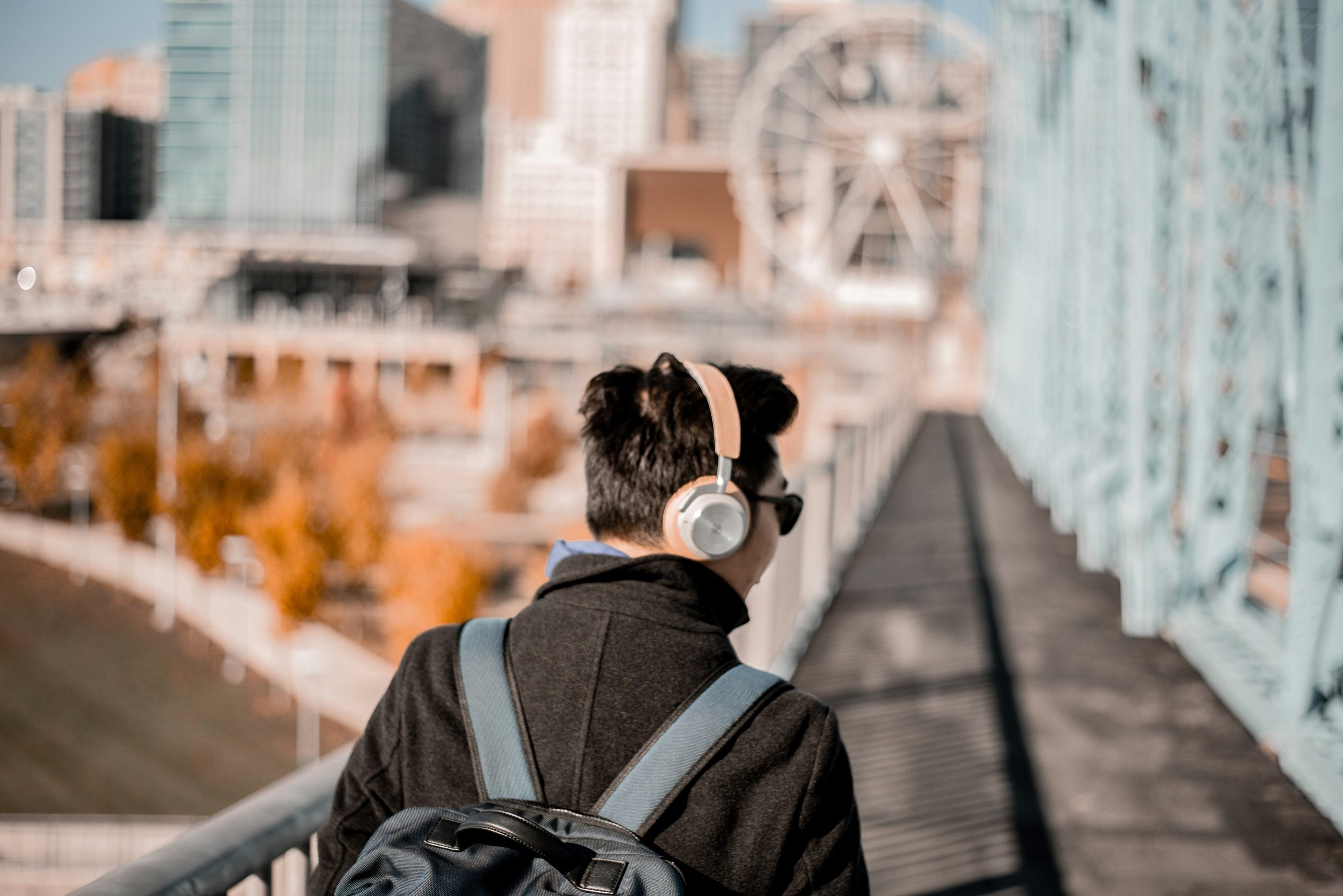 A man wearing wireless headphones and a backpack walks across a bridge with a city skyline and ferris wheel.