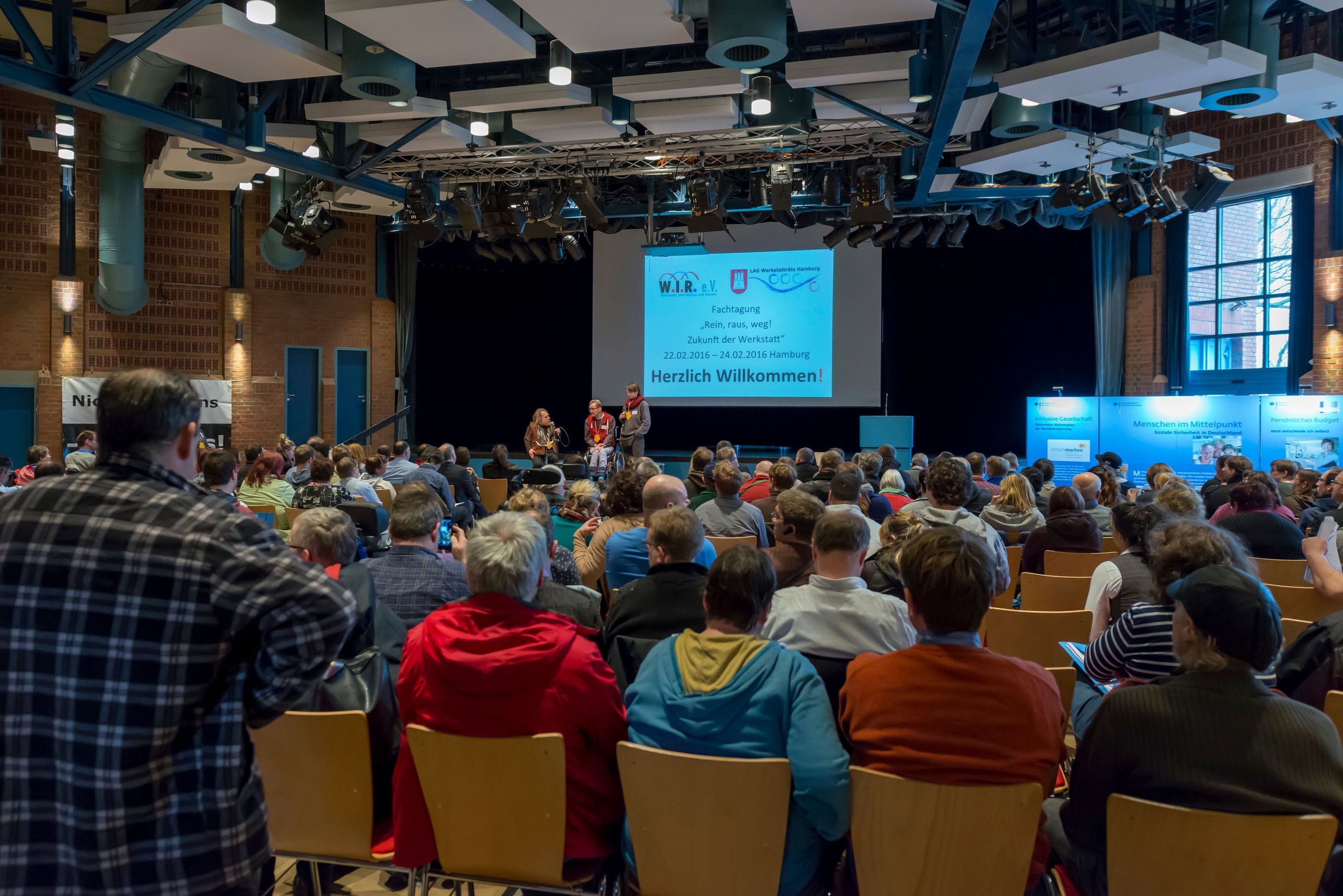 Viele Menschen sitzen im Publikum und hören den Redner*innen auf der Bühne zu.