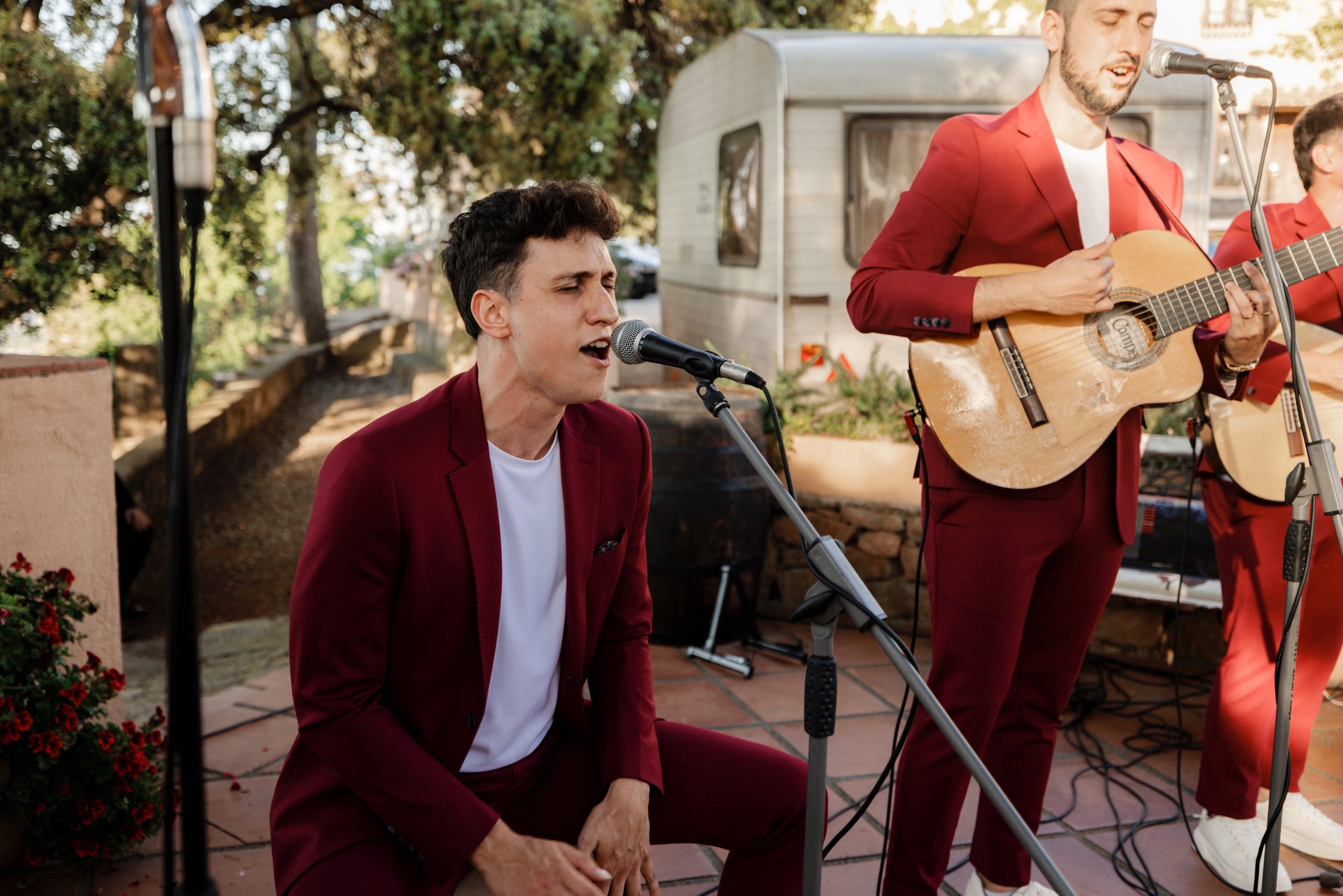 A live wedding band in burgundy suits performing outdoors with acoustic guitars and microphones.