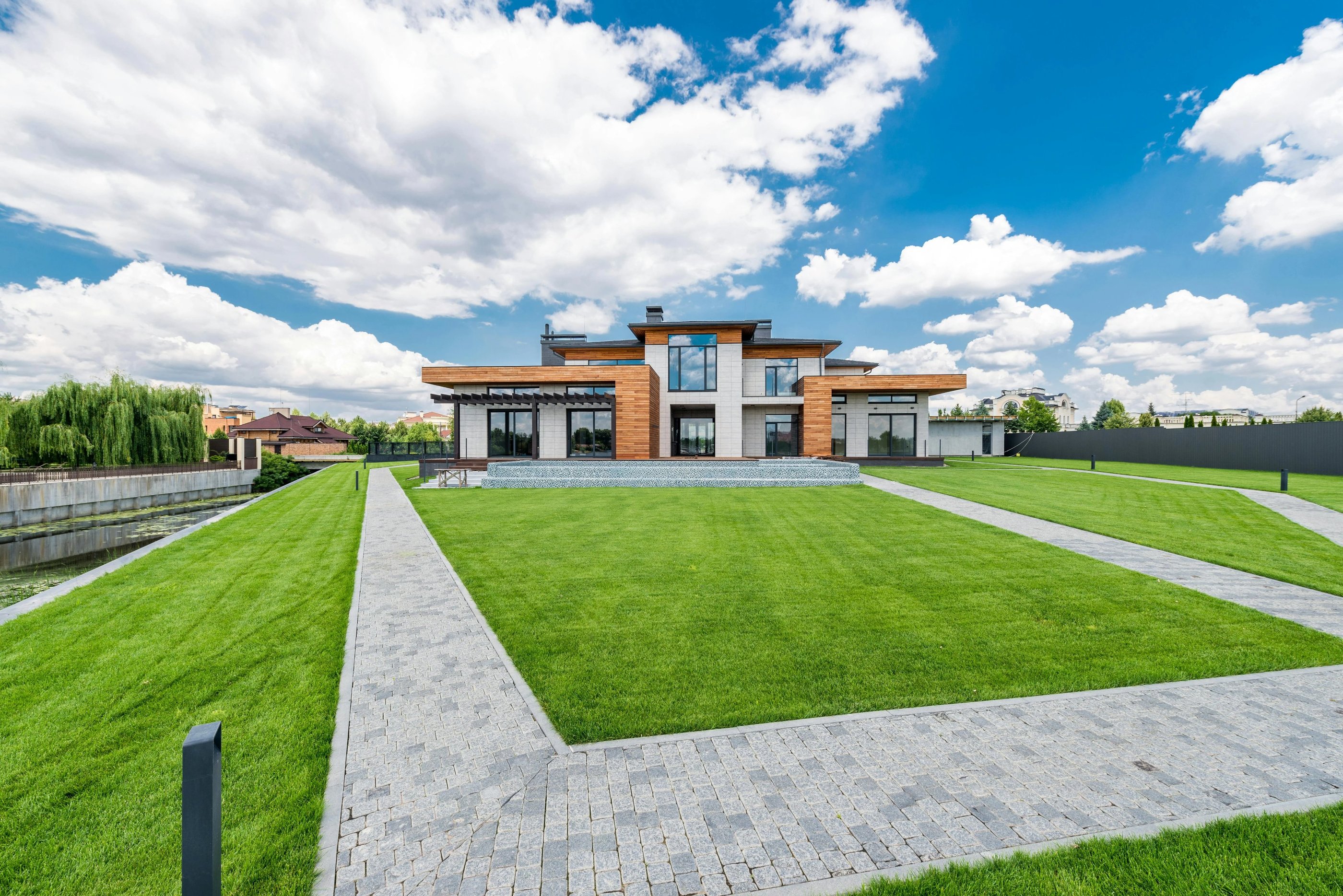 Modern luxury house with wood paneling, a large green lawn, and stone walkways under a blue sky.