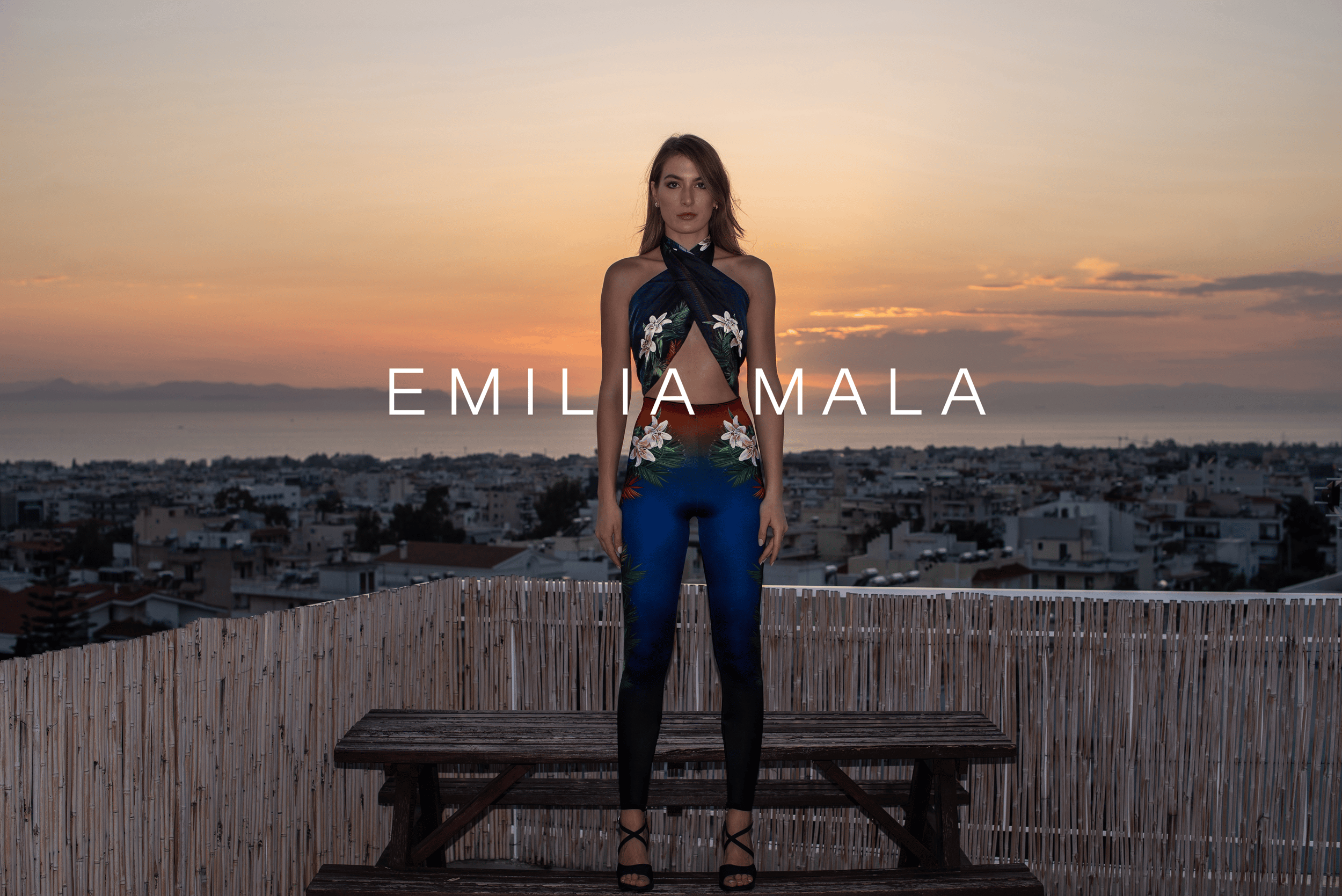 Model in Emilia Mala floral printed jumpsuit on a rooftop at sunset overlooking a city skyline.