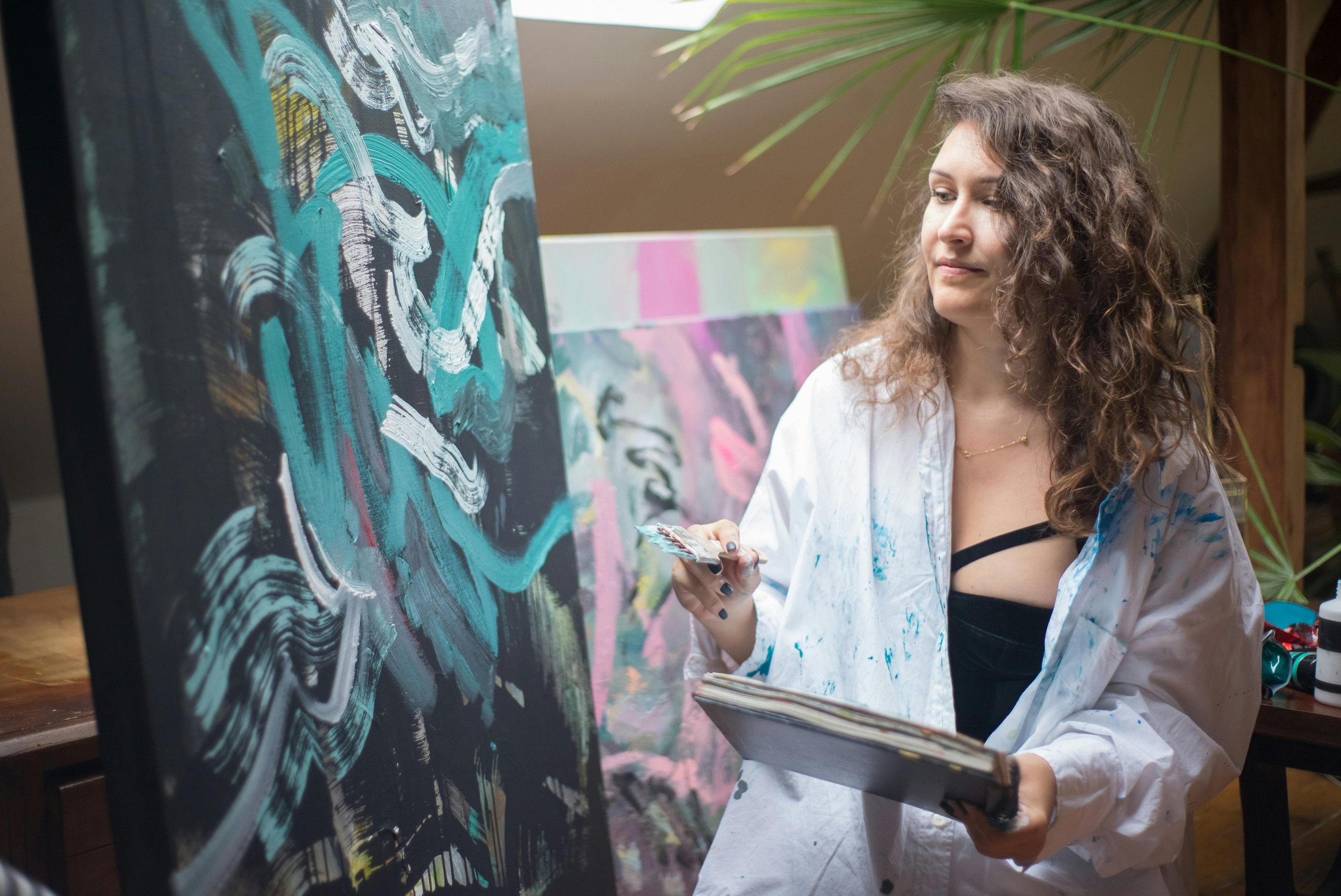 ovem mulher com camisa salpicada de tinta pintando em uma tela escura com pinceladas verde-azuladas.