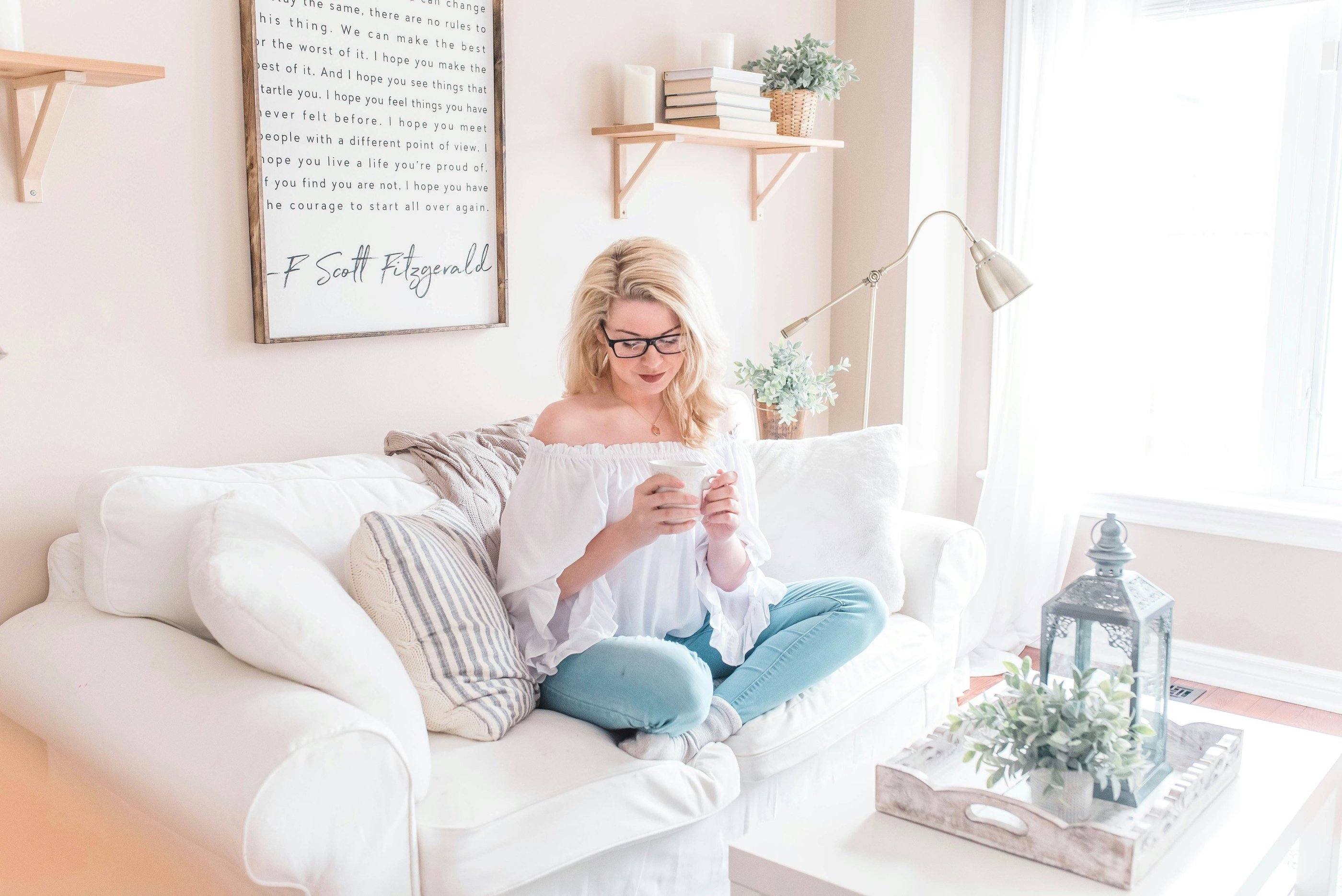 A blonde woman sitting on a white sofa in a bright living room, holding a mug.