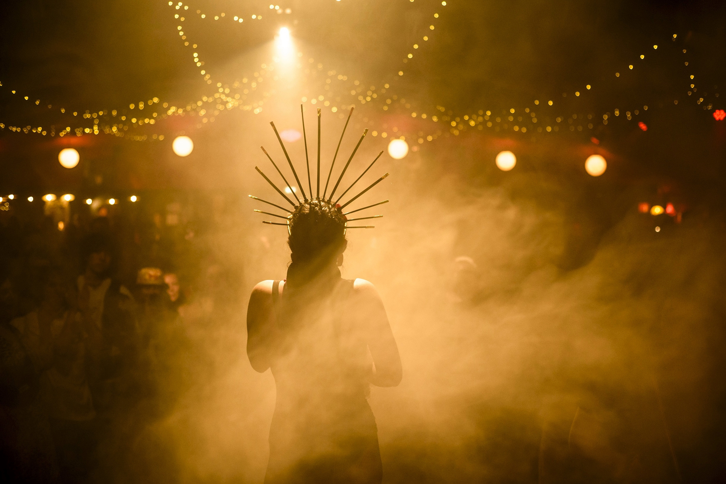 A performer wearing a gold spike crown standing in a hazy club with warm stage lighting and fairy lights.