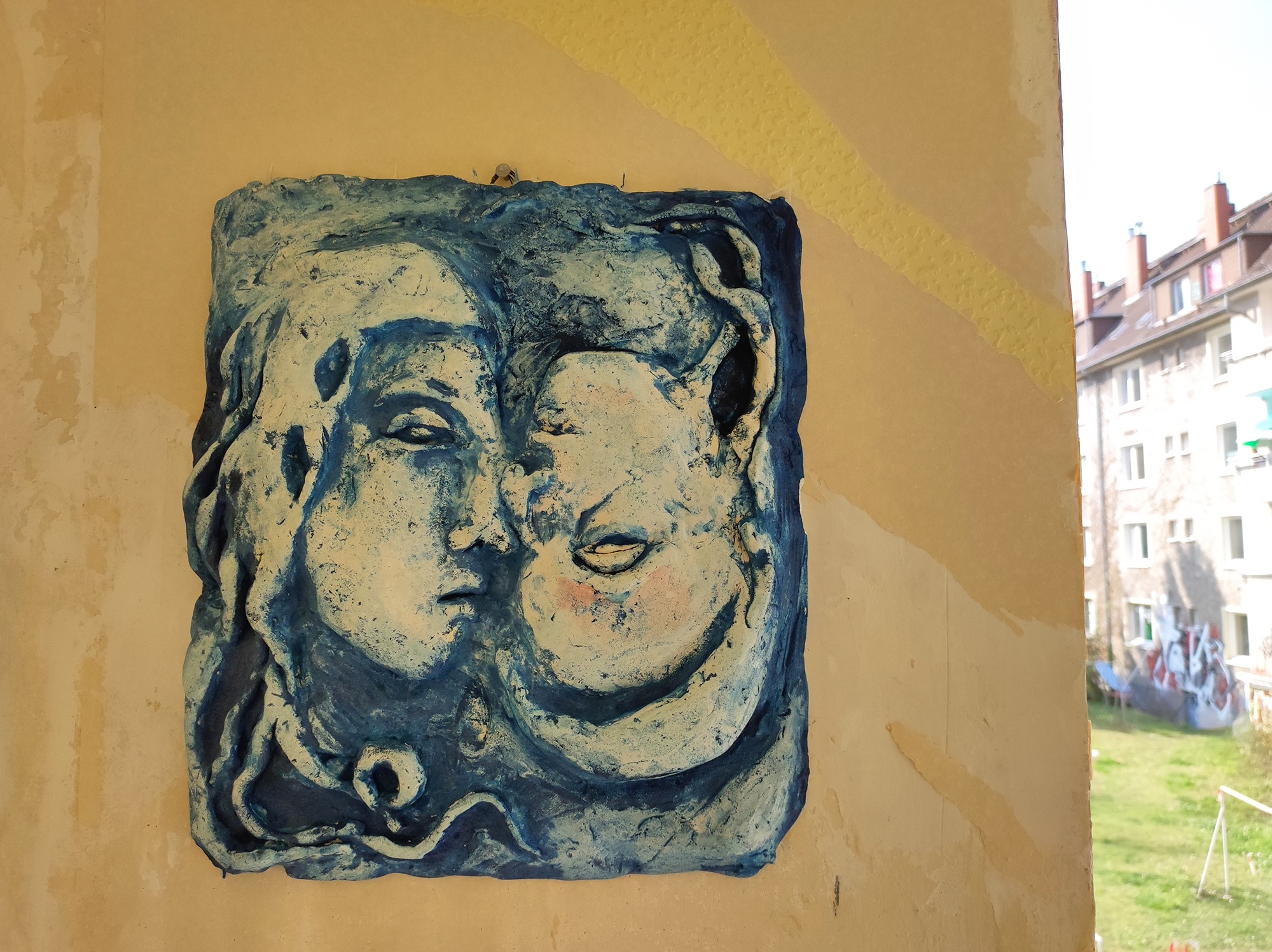 Blue ceramic wall sculpture depicting two faces in a textured, artistic relief on an exterior building wall.
