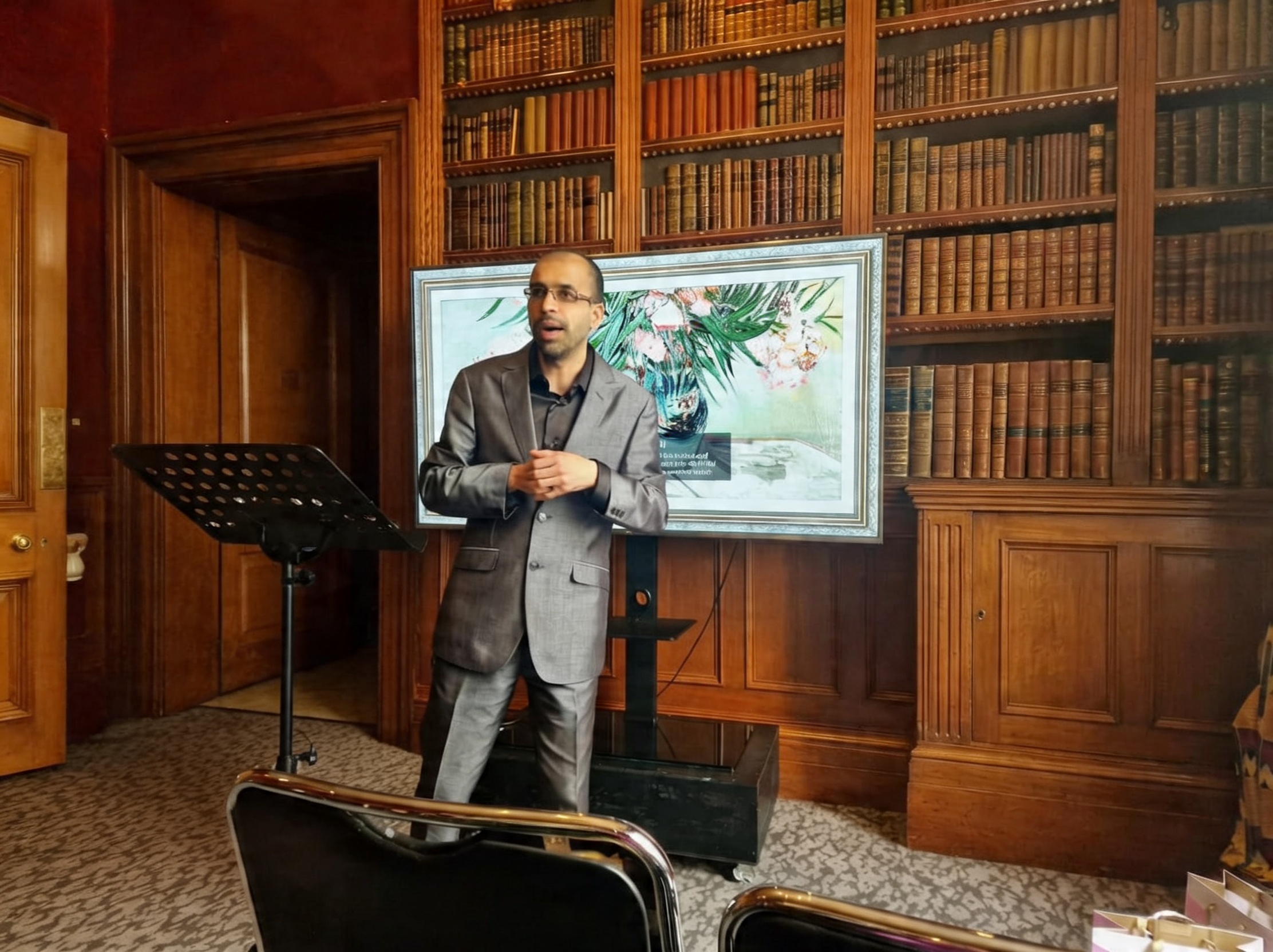 Professional speaker in a grey suit giving a presentation in a classic library with wood bookshelves.