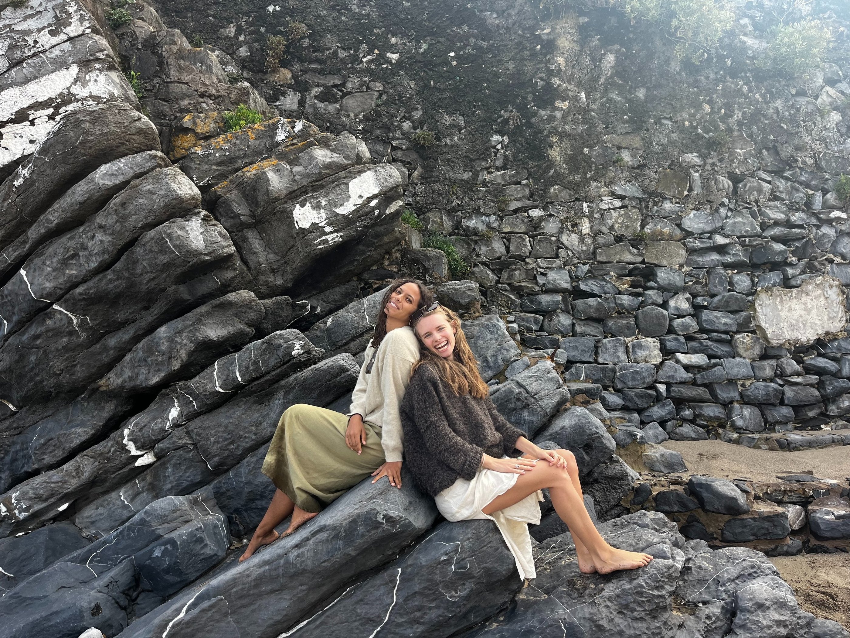 Two women sitting back to back on a rock.
