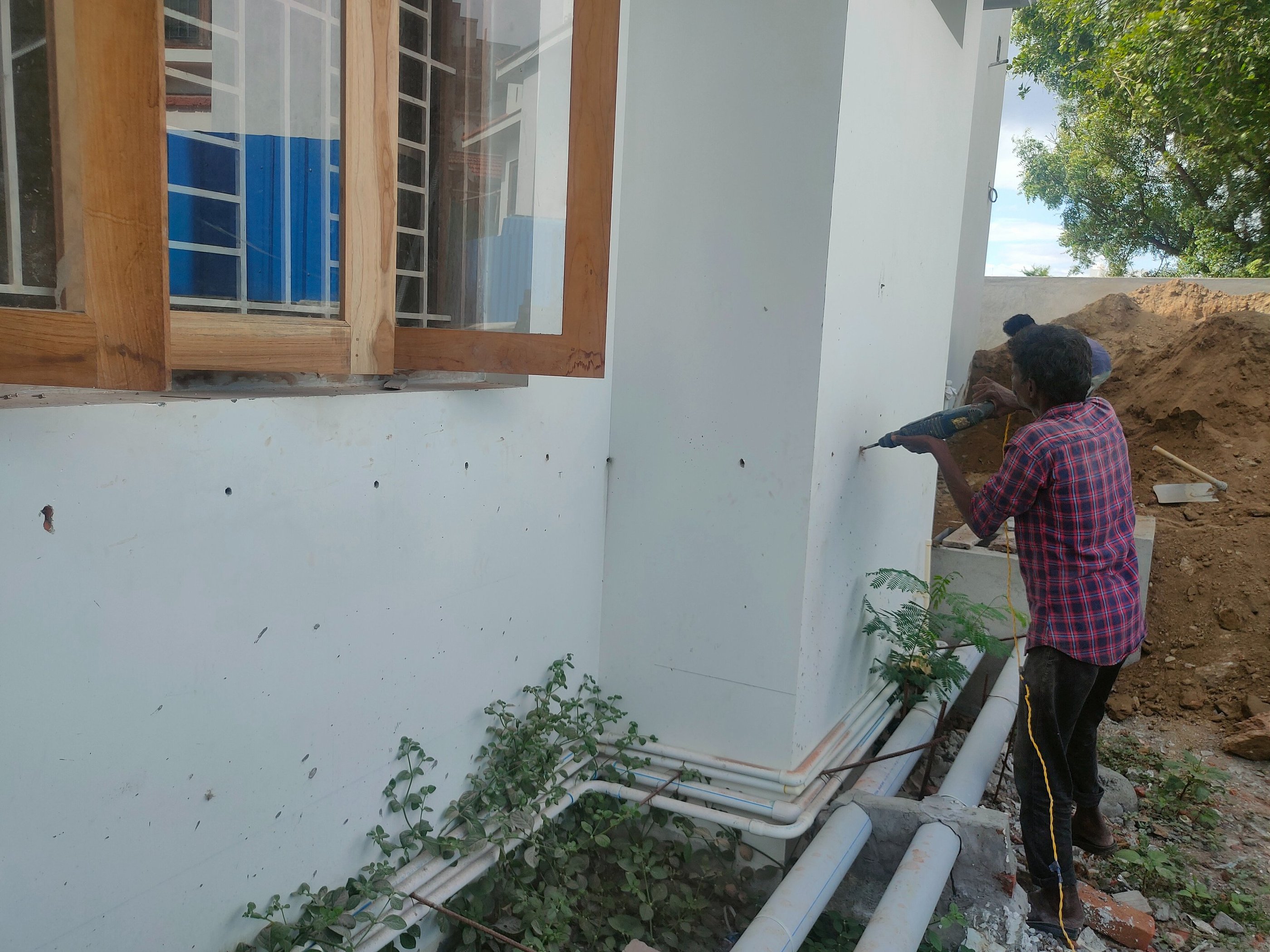 Professional pest control technician drilling exterior walls for termite treatment near a house window.