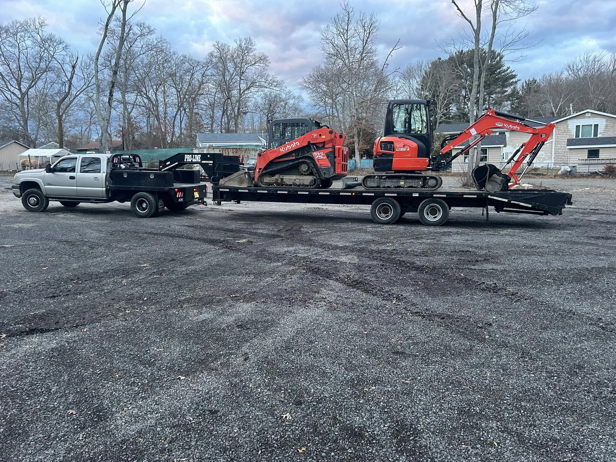 Flatbed truck towing a trailer with a Kubota skid steer and mini excavator on a gravel lot.