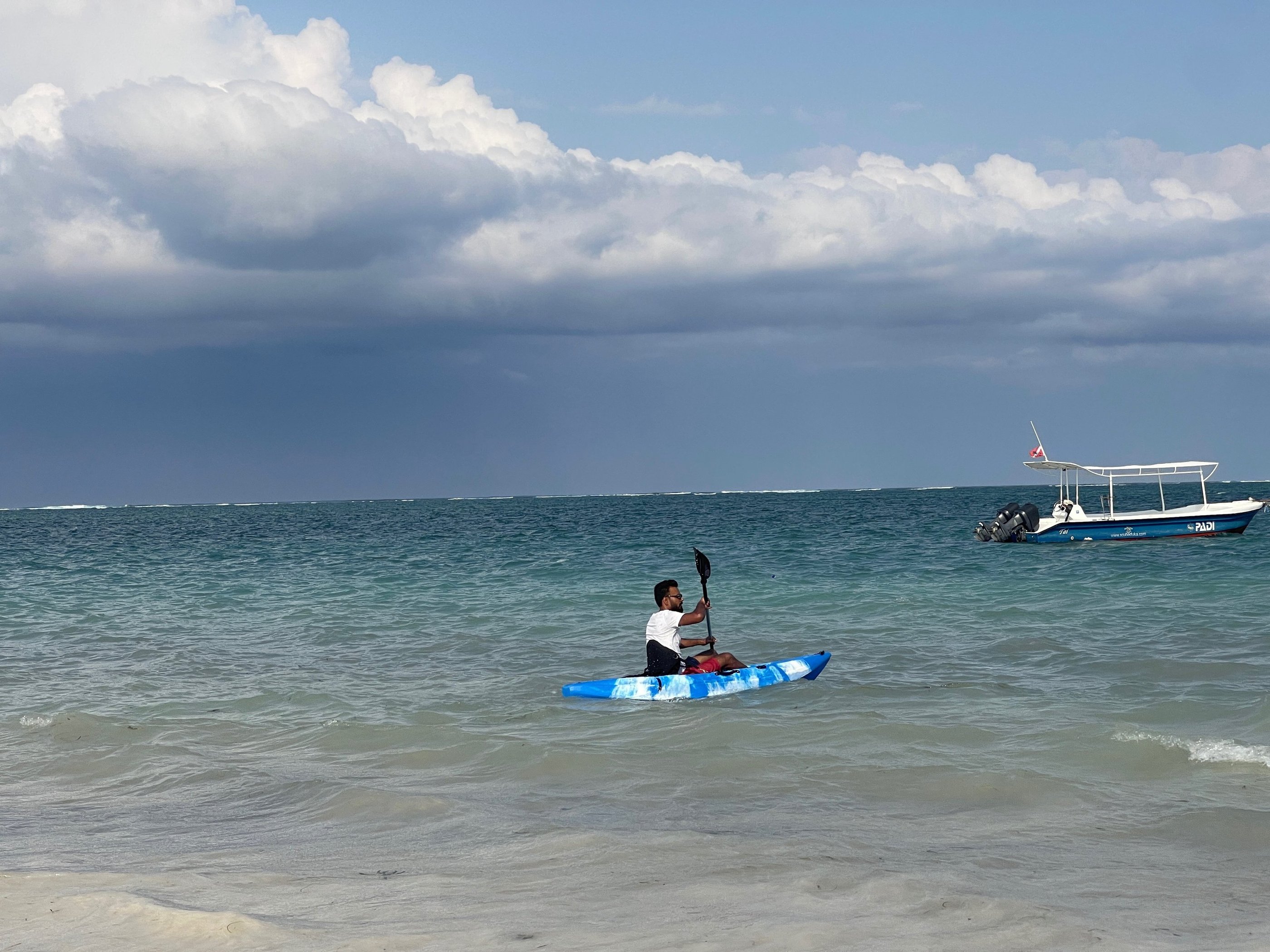 A man paddles a blue camouflage kayak in tropical turquoise ocean waters near a white boat.