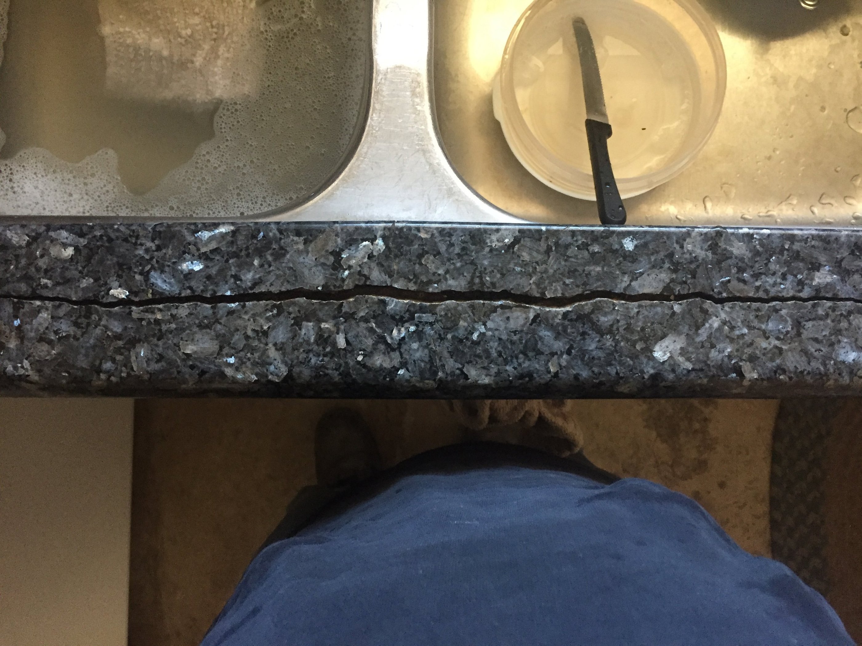 A long horizontal crack in a dark grey granite kitchen countertop next to a stainless steel sink.