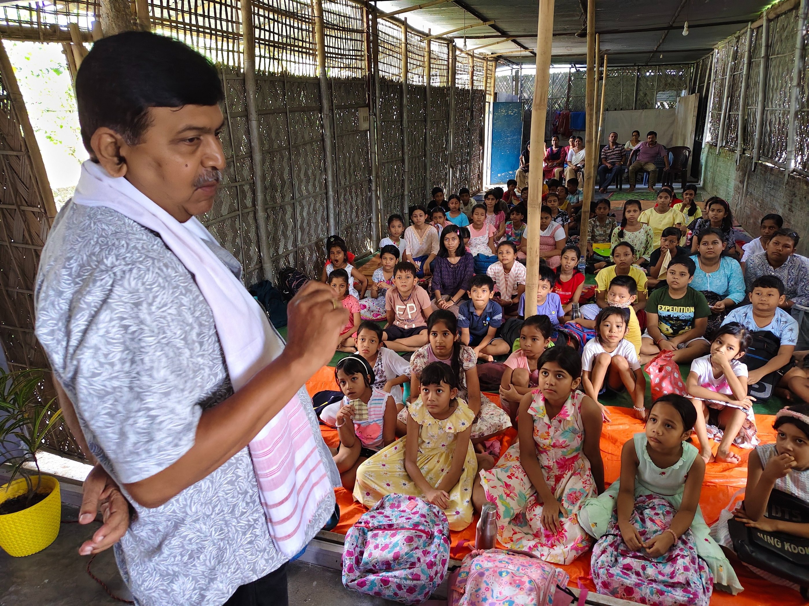 An educator teaching a large group of attentive children sitting on the floor in a bamboo-walled classroom.