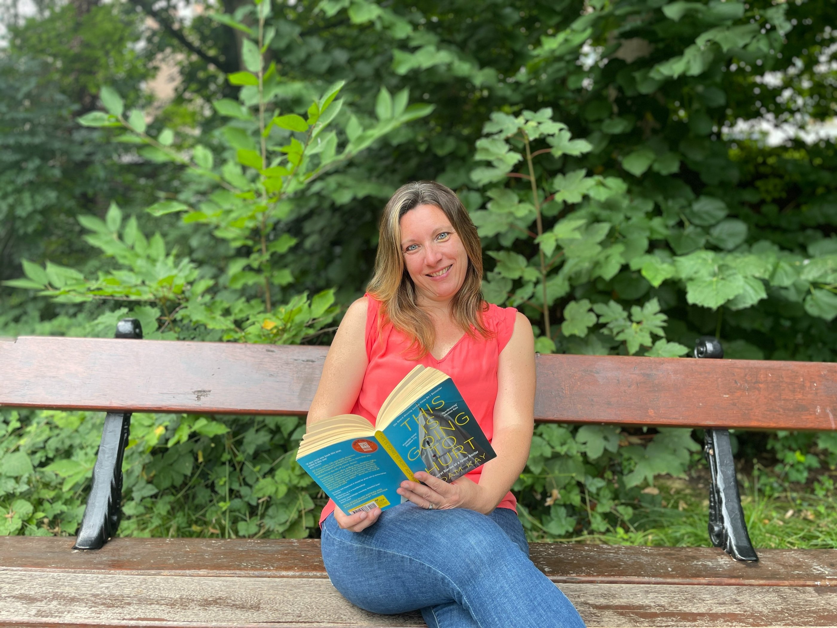 Portrait photo of Jen Ruthe sitting on a park bench in a red top, holding a book.