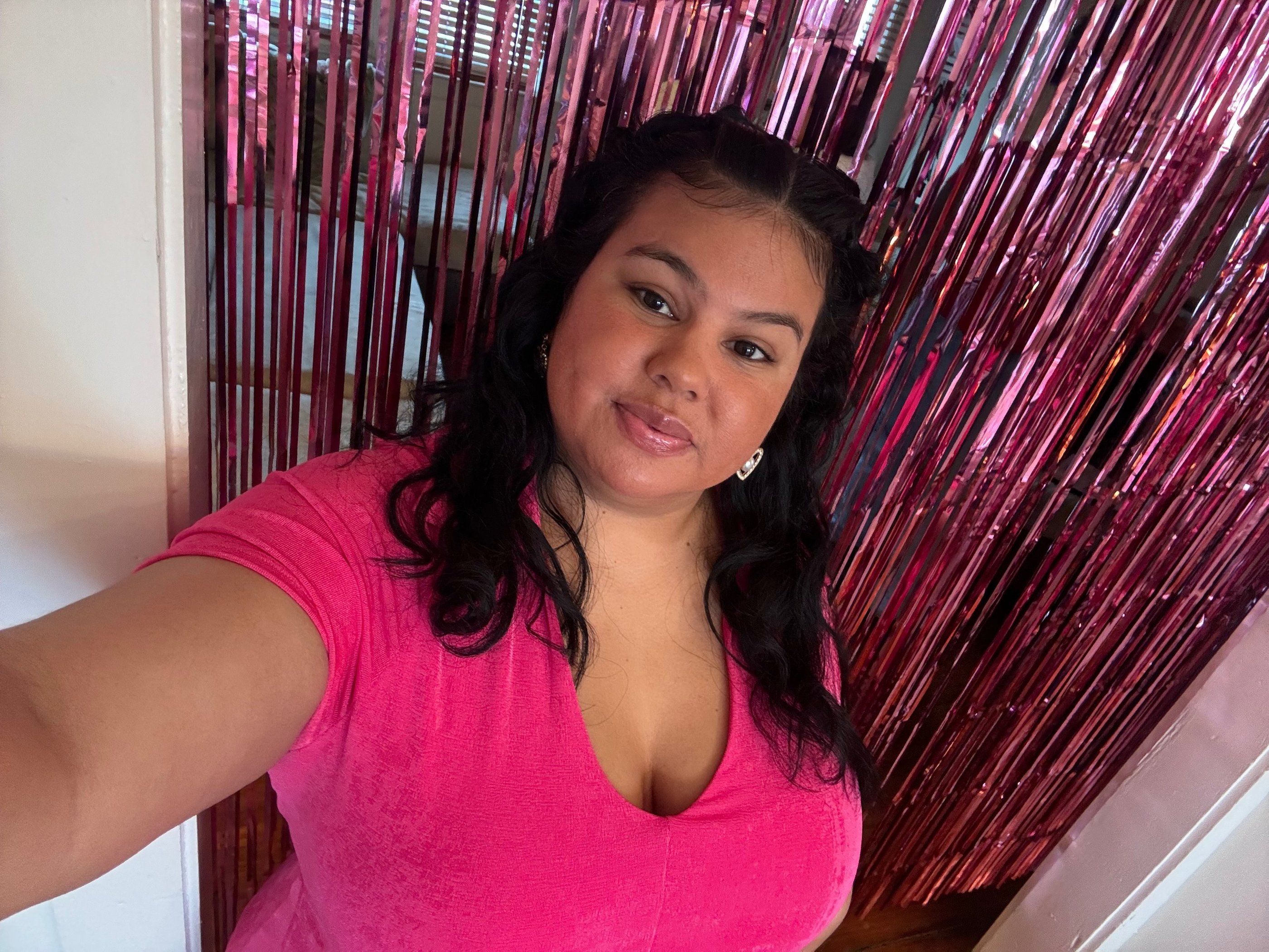 A smiling woman in a pink t-shirt poses in front of a shiny pink foil fringe backdrop.
