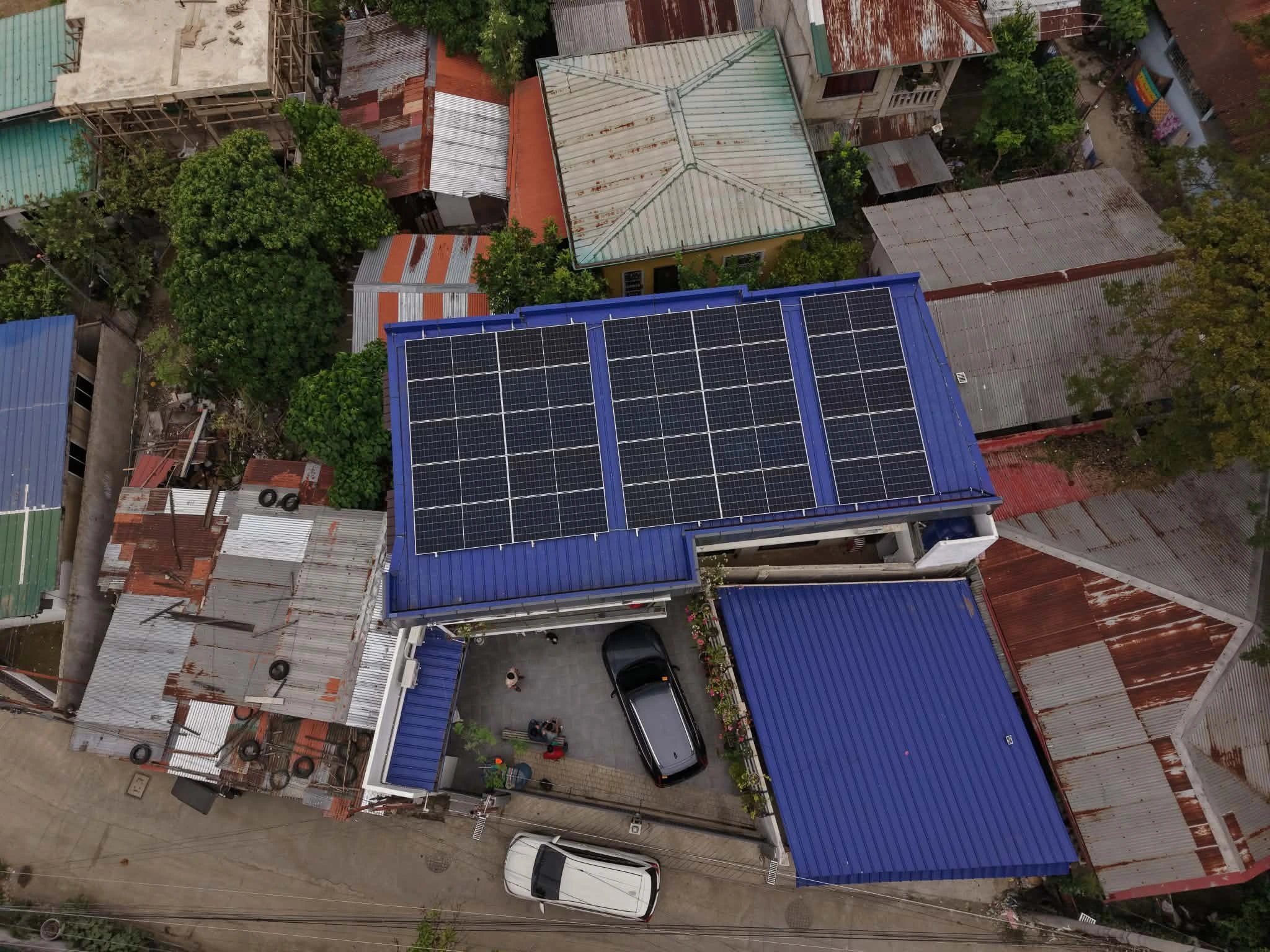 An aerial drone shot of a residential house with a bright blue metal roof featuring a large array of black solar panels