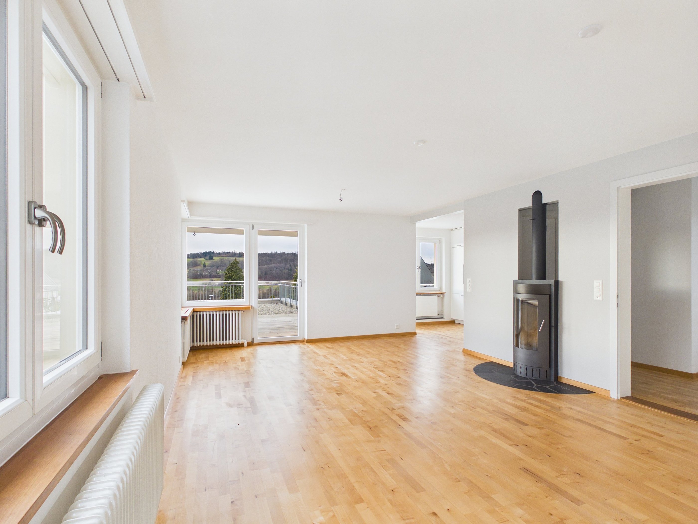 Empty modern living room featuring light hardwood floors, a black wood-burning stove, and large glass balcony doors.