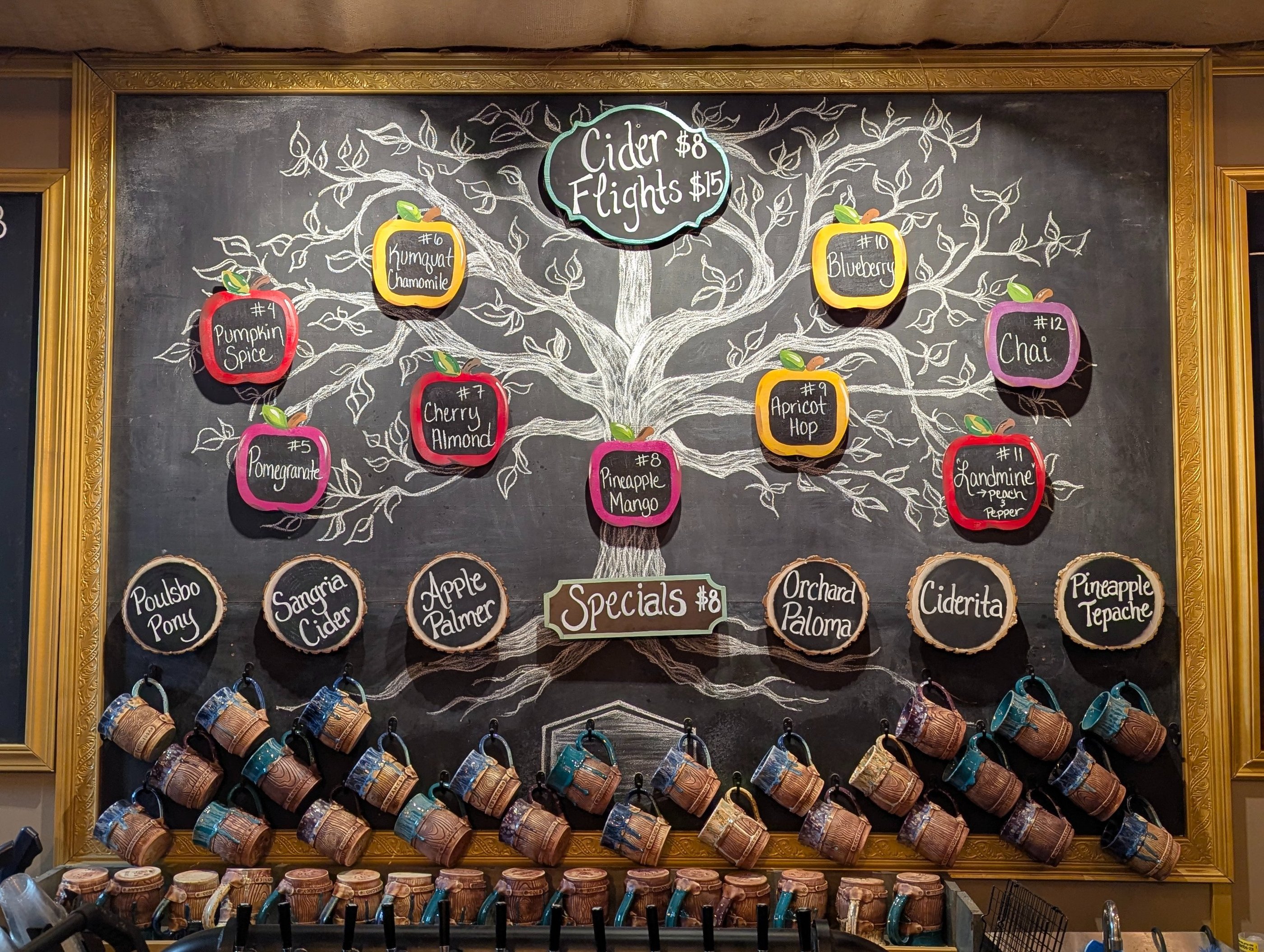 Hand-drawn chalkboard menu featuring hard cider flight flavors and specials above a row of ceramic mugs.