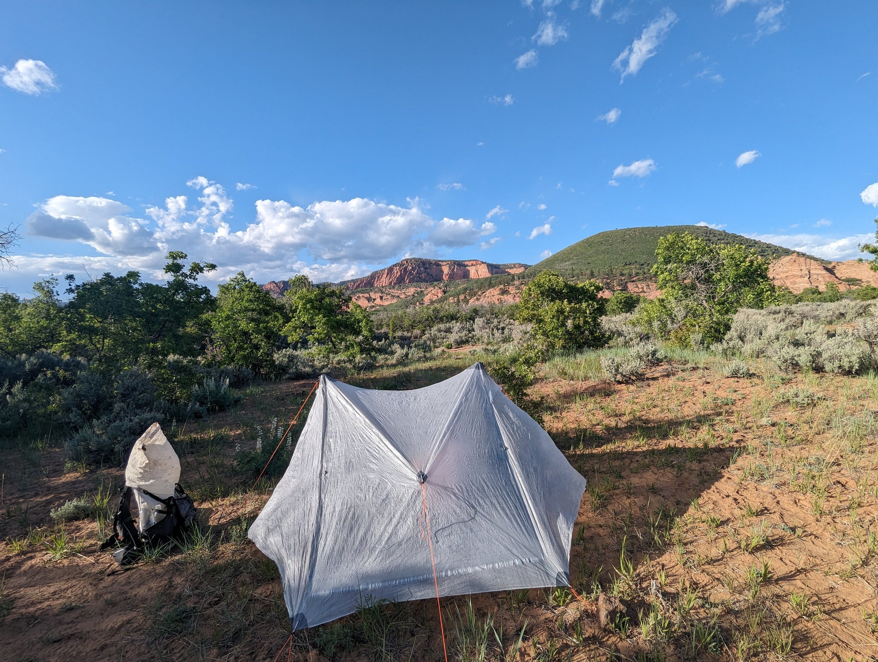 A backpacking tent pitched in a desert wilderness camp with red rock mountains under a blue sky.