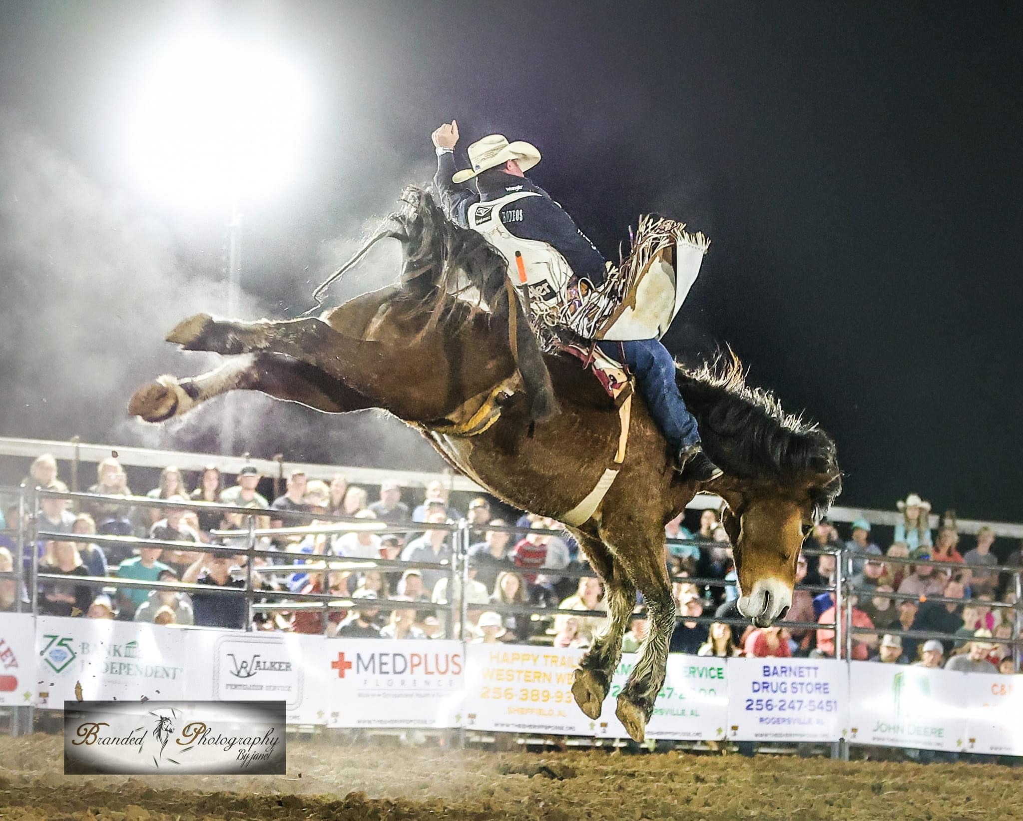 A professional cowboy competing in a bareback bronc riding event at a night rodeo arena.