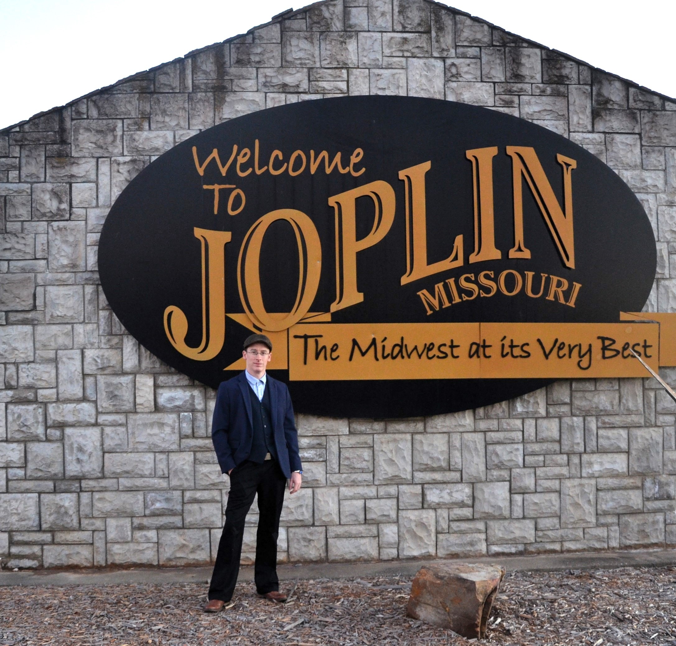A man in a suit standing in front of the Welcome to Joplin Missouri sign on a stone wall.