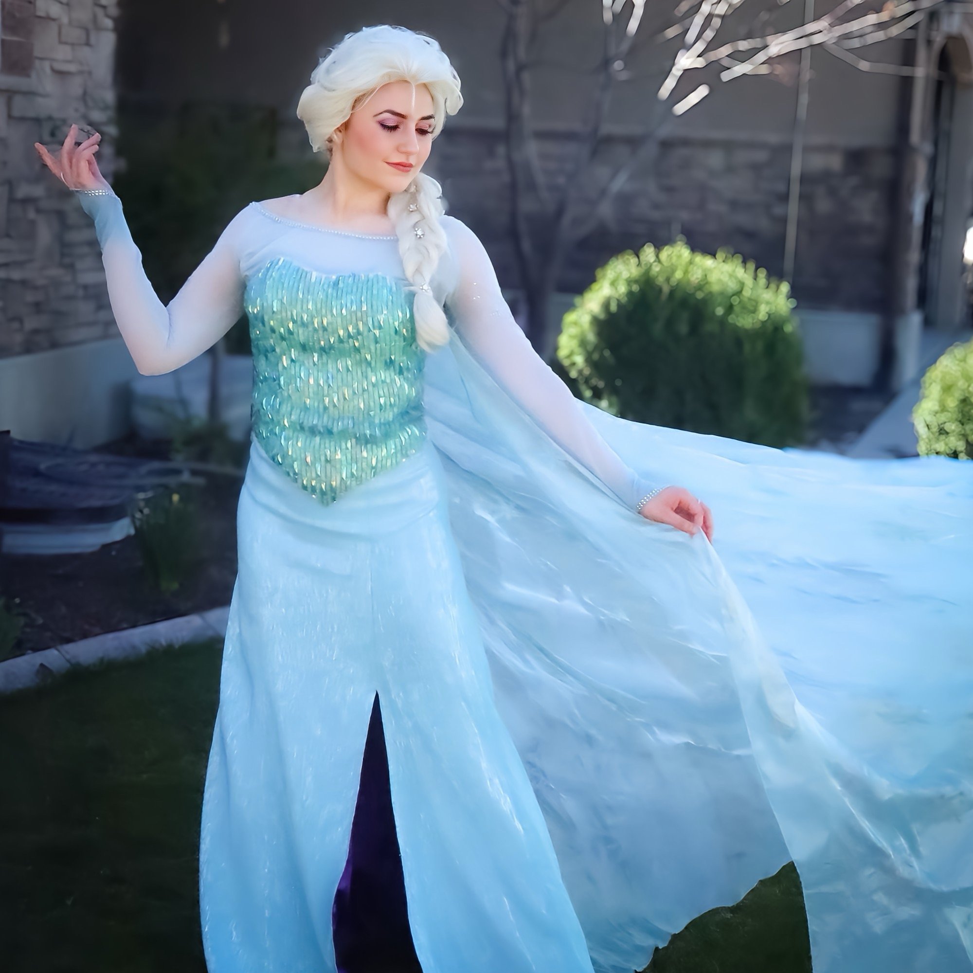 A woman in a sparkling Elsa Frozen cosplay costume with a blonde braid posing outdoors.