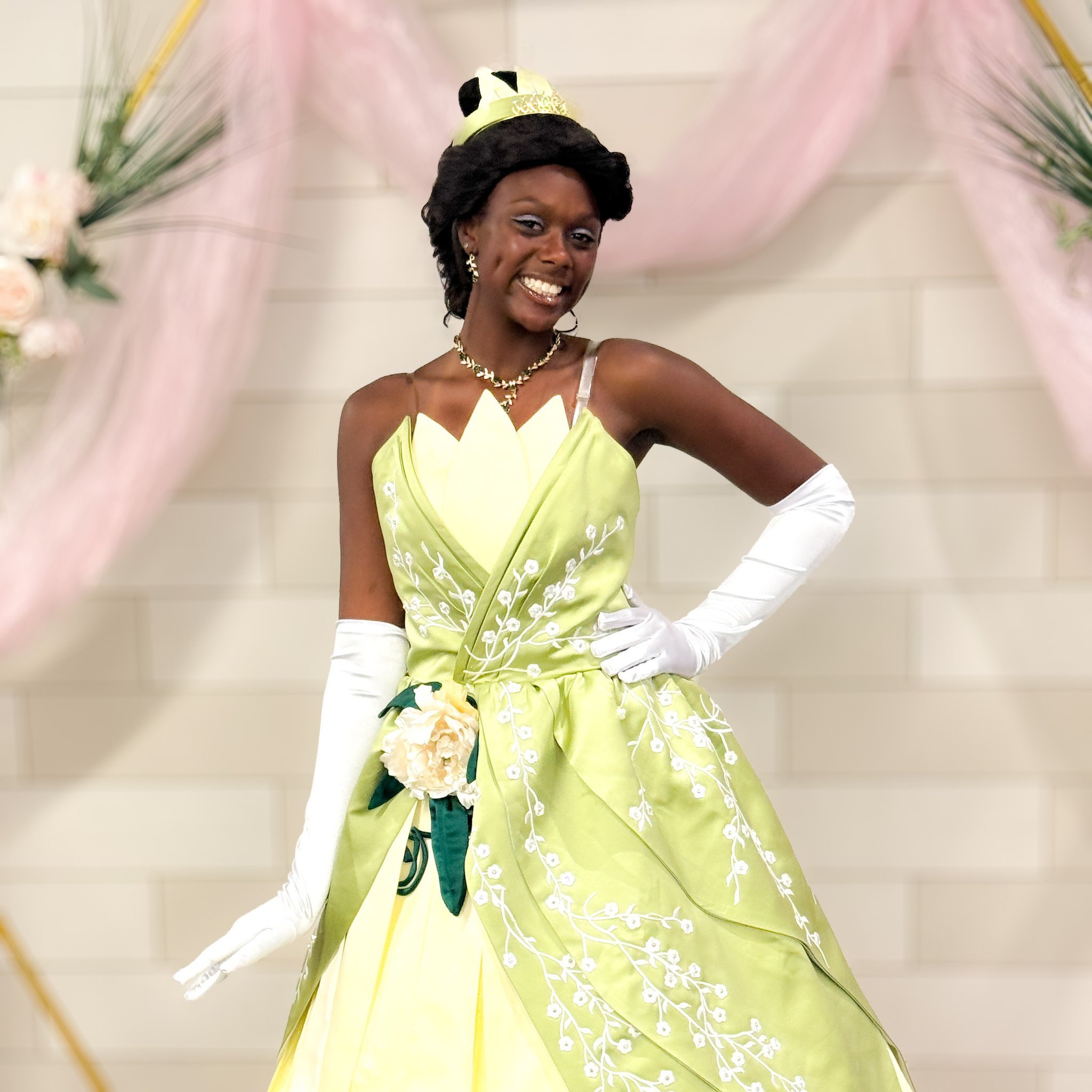 Smiling woman in a green Princess Tiana cosplay dress and white gloves posing against a floral backdrop.