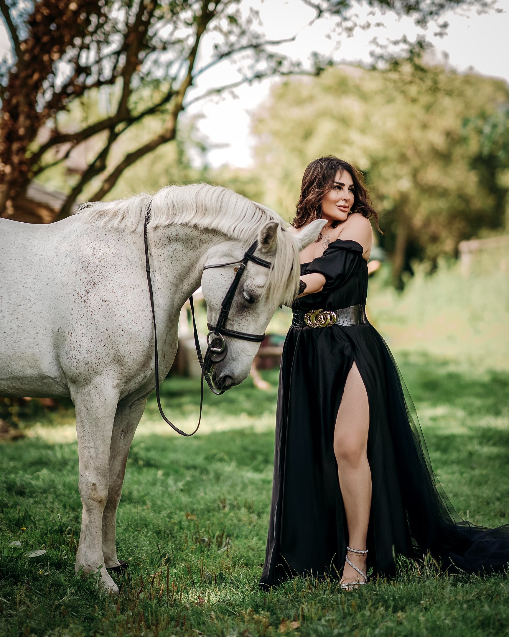 Model in black dress posing beside a white horse in nature, photographed by Fred Art Studio.