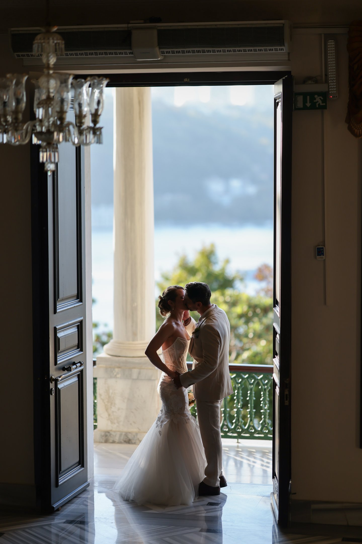 Newlyweds kiss on a balcony overlooking water, framed by open doors Elaine Brackin, Kenan Kasap