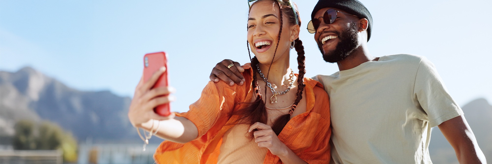 A smiling young couple takes a selfie on a red smartphone with mountains in the background.
