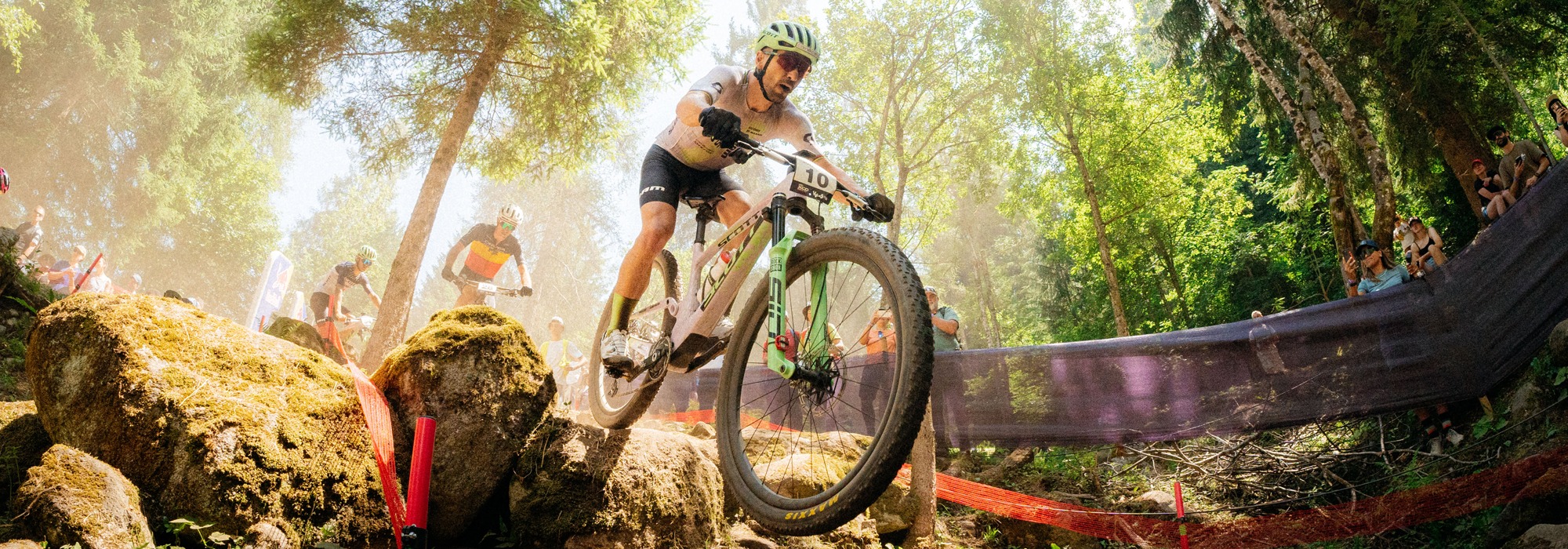 Professional mountain biker racing down a rocky forest trail during a cross-country MTB competition.
