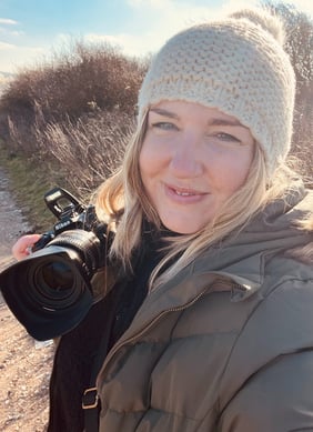 A profile photo of Claire Walker, smiling holding her Nikon D7500. Shes wearing a green winter coat and a create beanie hat. 