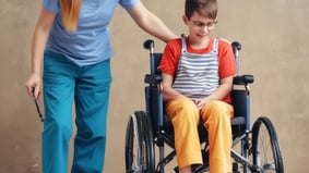 A woman assists a boy in a wheelchair, providing support and guidance in a caring manner.