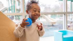 Smiling child with Down syndrome playing indoors with a blue ball near window